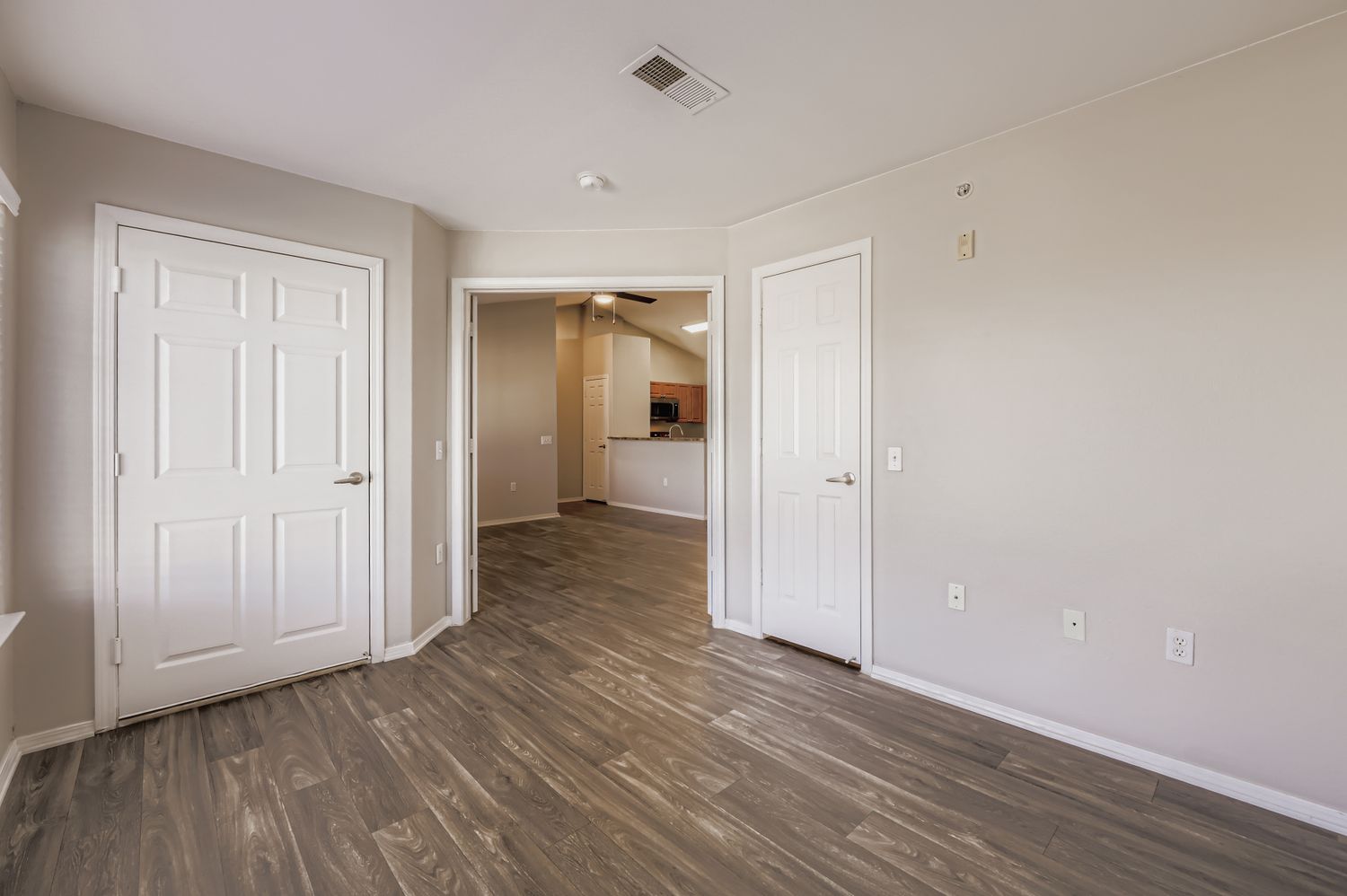 A spacious, light-filled room featuring neutral walls, a door with a silver handle, and wide-plank laminate flooring. Two doorways lead to adjacent areas, with the one on the right opening into a kitchen space. The layout offers a modern and inviting atmosphere suitable for various uses.