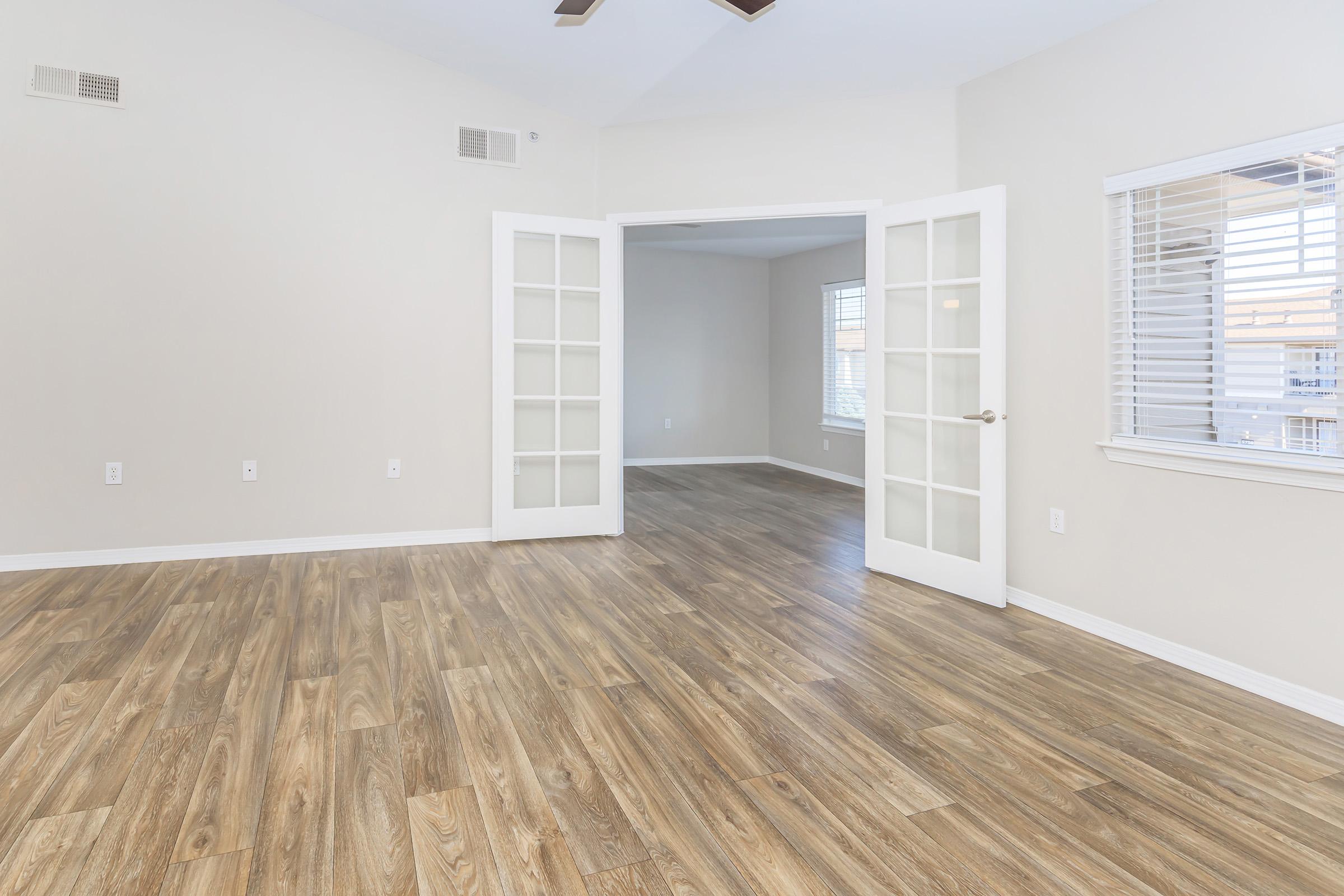 Spacious room with light-colored walls and hardwood flooring, featuring double French doors leading to another room. Natural light streams in through a window with blinds. The ceiling is high with a fan. The room appears clean and modern, suitable for various uses.