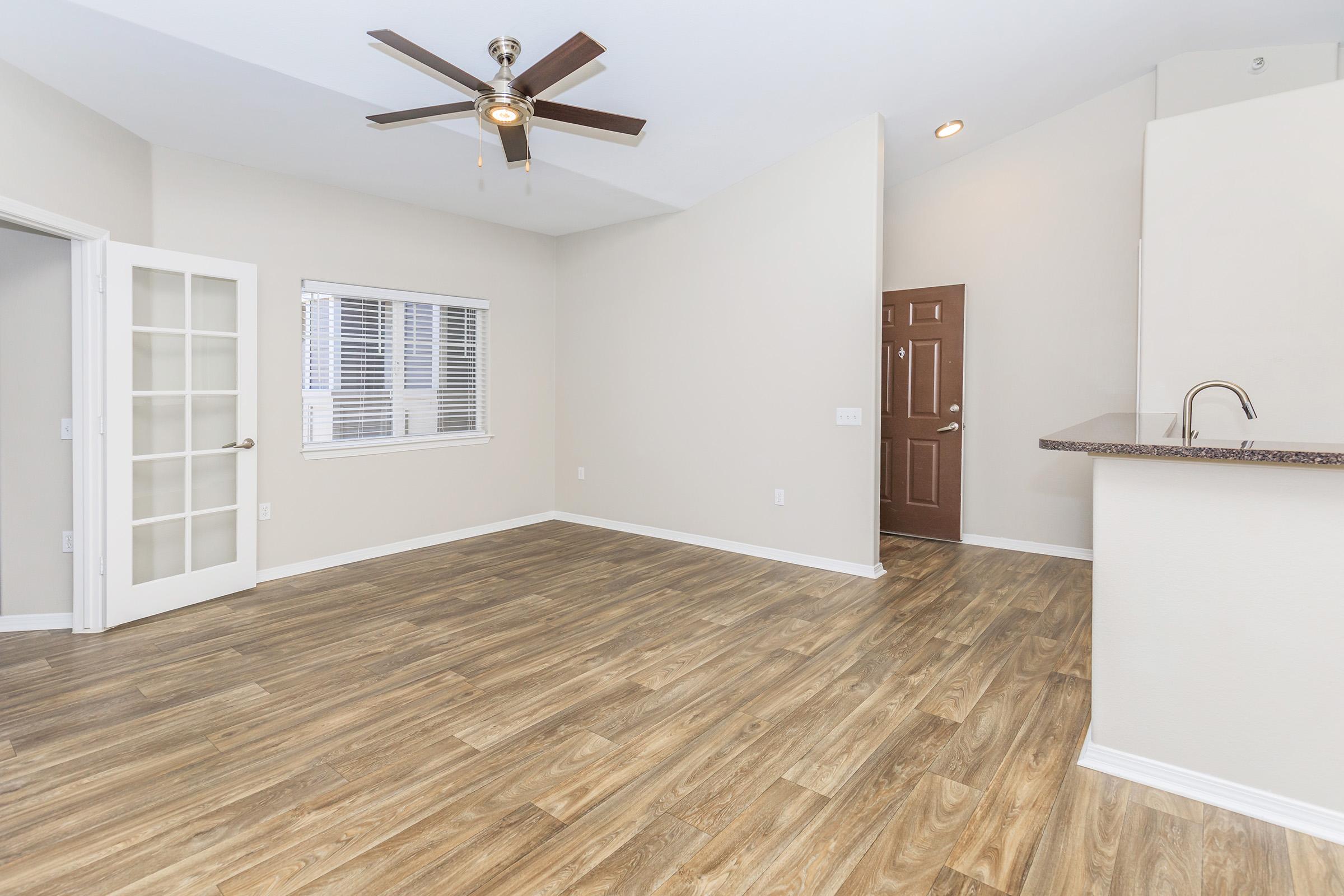 Bright, open living space with light wood flooring, a ceiling fan, and neutral-colored walls. A large window allows natural light in, and there are glass French doors leading to another room. The kitchen area features a bar counter, and the front door is to the right. Ideal for a modern apartment or home.