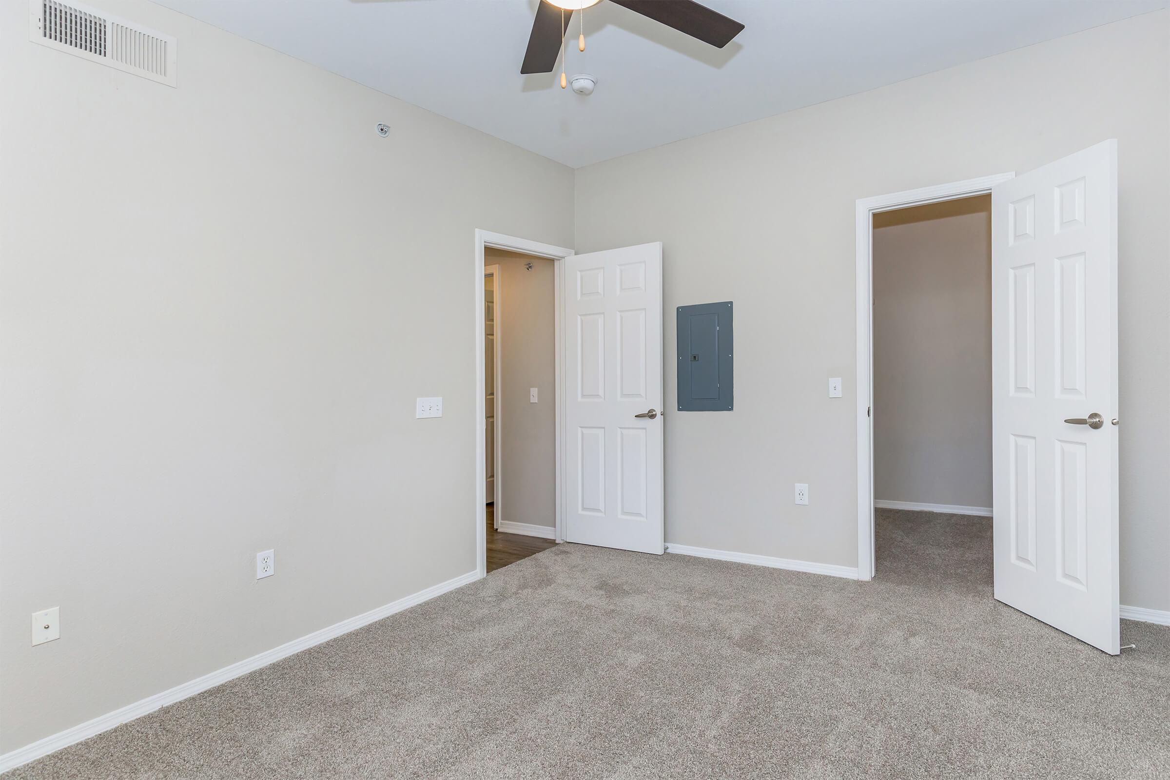 A spacious room with light beige walls and plush carpeting. There are two white doors on the left and right, and a third door partially open in the back. A ceiling fan is visible above, providing a modern touch to the simple interior. The room is well-lit and appears ready for furnishing.