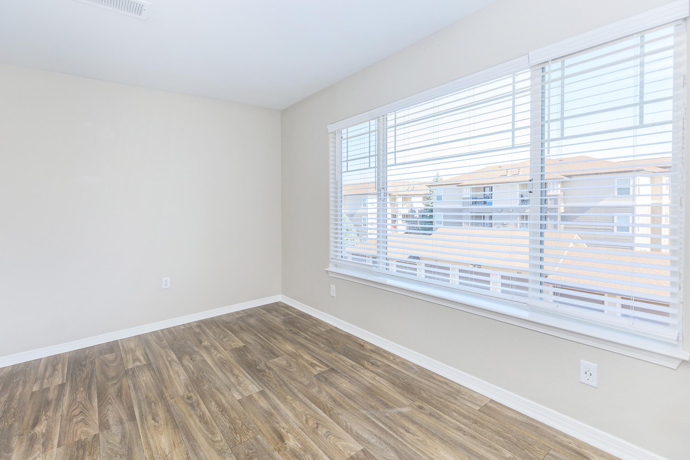Bright and spacious room with light-colored walls and wooden flooring. It features a large window with horizontal blinds, allowing natural light to fill the space. The view outside includes neighboring buildings, creating a welcoming atmosphere suitable for various uses.