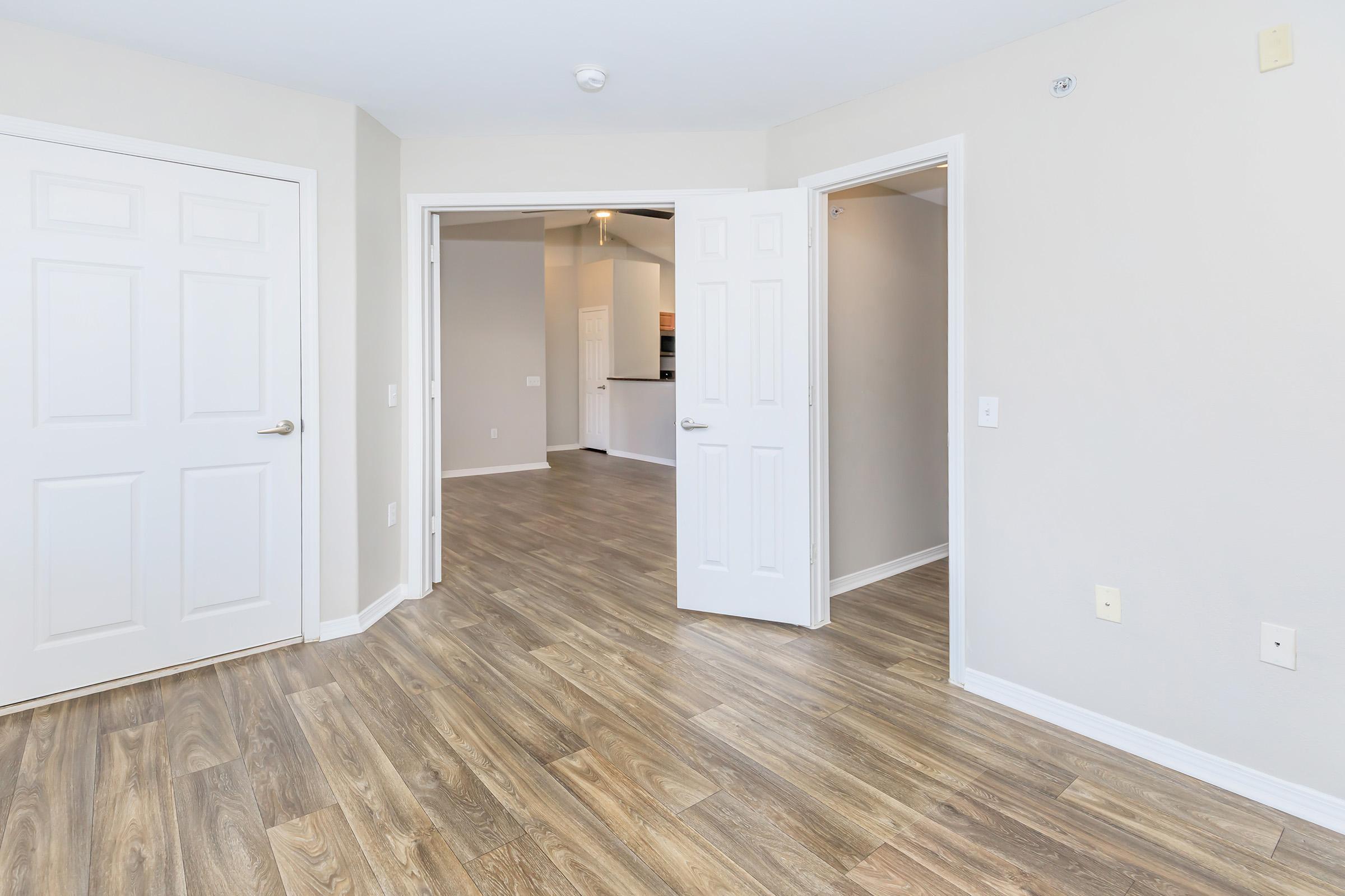 An interior view of a room featuring light-colored walls and wood-like flooring. Two doors are visible, leading to adjacent spaces. The room appears spacious and well-lit, with a doorway in the background leading to a kitchen area.