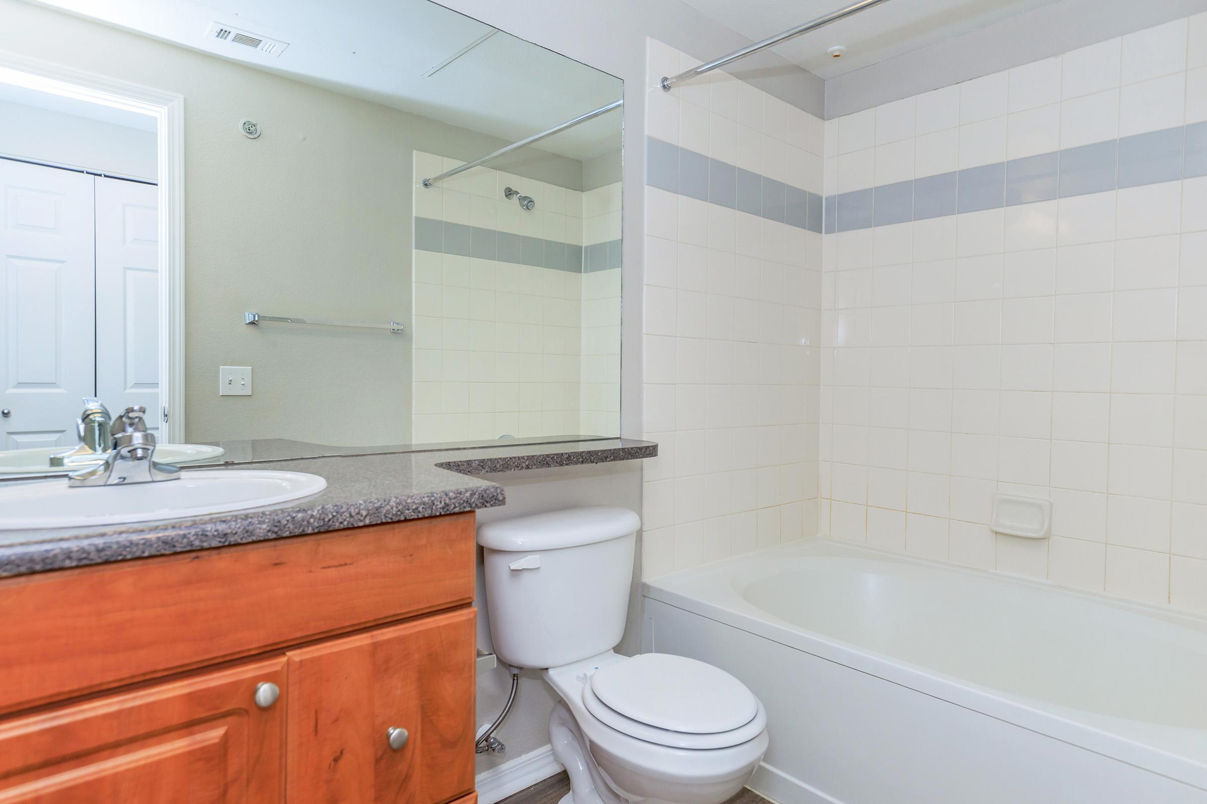 A modern bathroom featuring a white bathtub with a shower, a wall-mounted mirror above a sink with a dark countertop, and a wooden vanity. The walls are tiled in light colors, and there's a glimpse of closed double doors in the background.