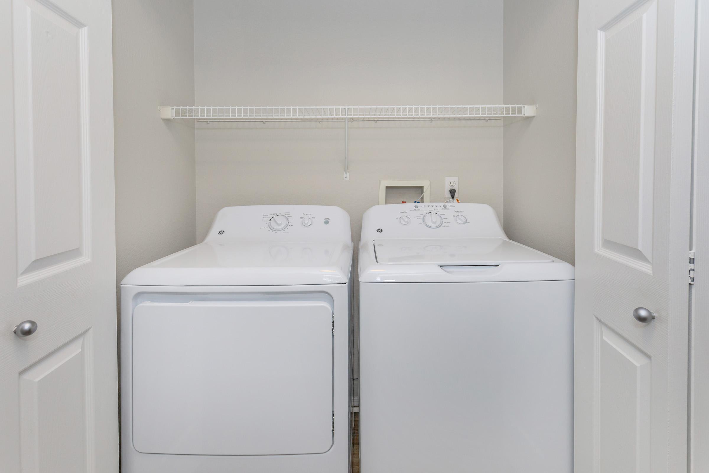 A laundry room featuring a white washer and dryer side by side, with a shelf above them. The walls are light-colored, and there are closed double doors in the foreground. The space is clean and organized, suitable for household laundry tasks.