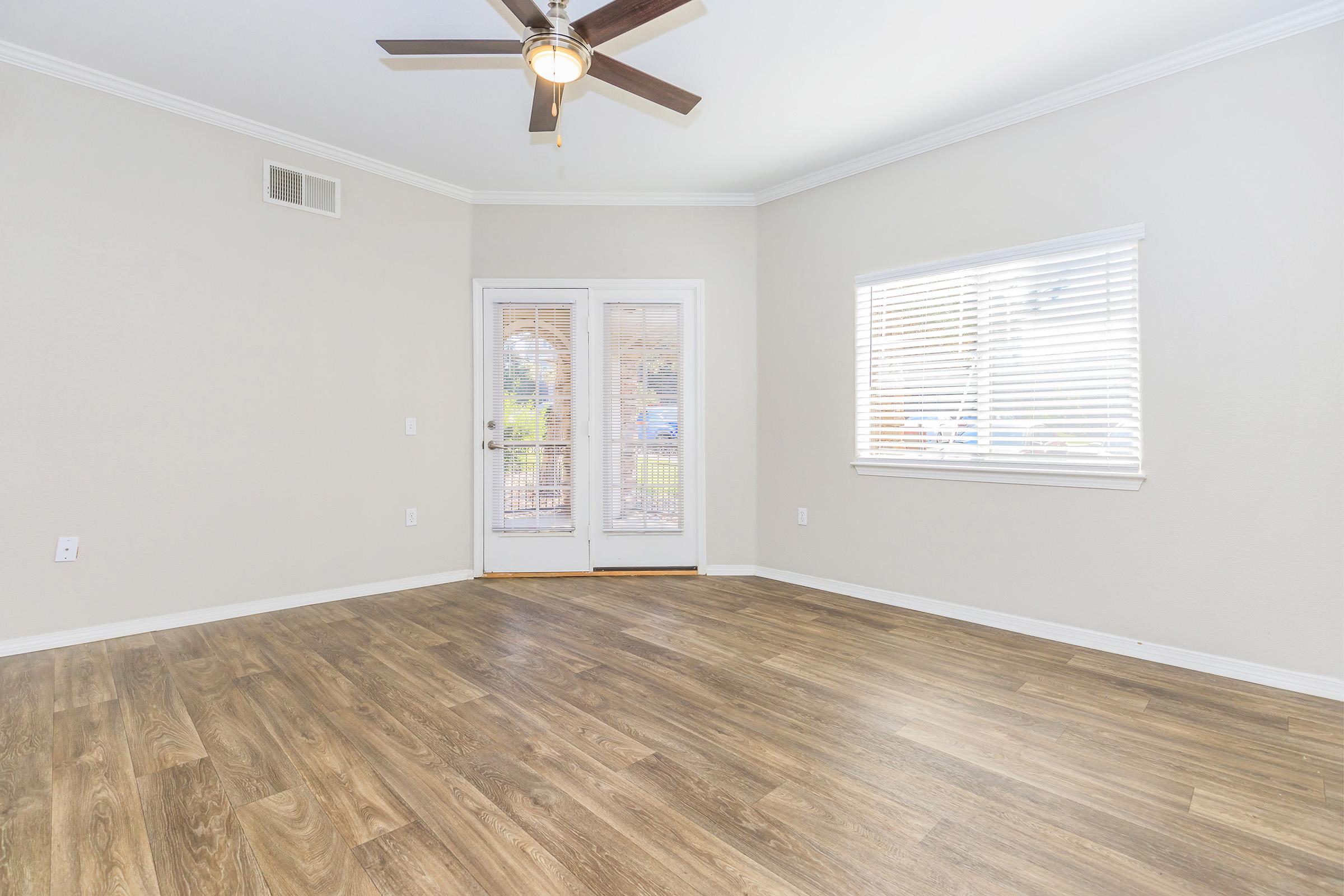 A spacious, empty room featuring light-colored walls, a ceiling fan, and laminate flooring. There are double glass doors leading to an outdoor area, and a window with blinds allowing natural light to enter. The room has a clean, modern aesthetic.