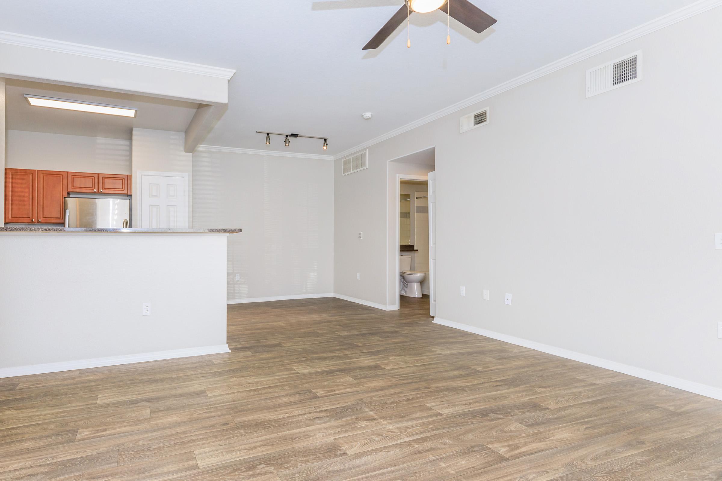 Spacious interior of an apartment featuring a living area with wood-style flooring, ceiling fan, and open kitchen with wooden cabinets. Natural light fills the room, and a hallway leads to a bathroom in the background.
