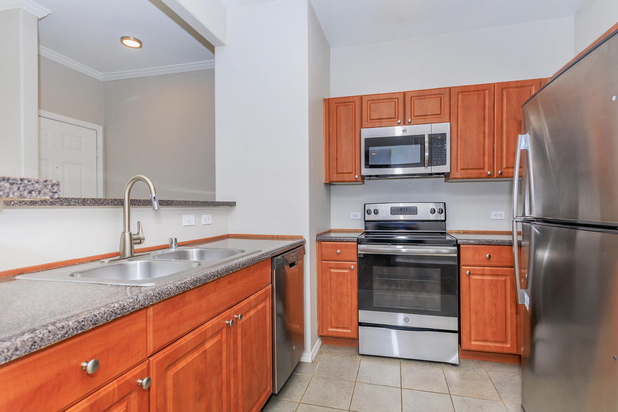 View of a modern kitchen featuring wooden cabinetry, a stainless steel refrigerator, a microwave, and an oven. The kitchen has a double sink, granite countertops, and tiled flooring, providing a clean and functional cooking space. Bright lighting enhances the overall welcoming atmosphere.