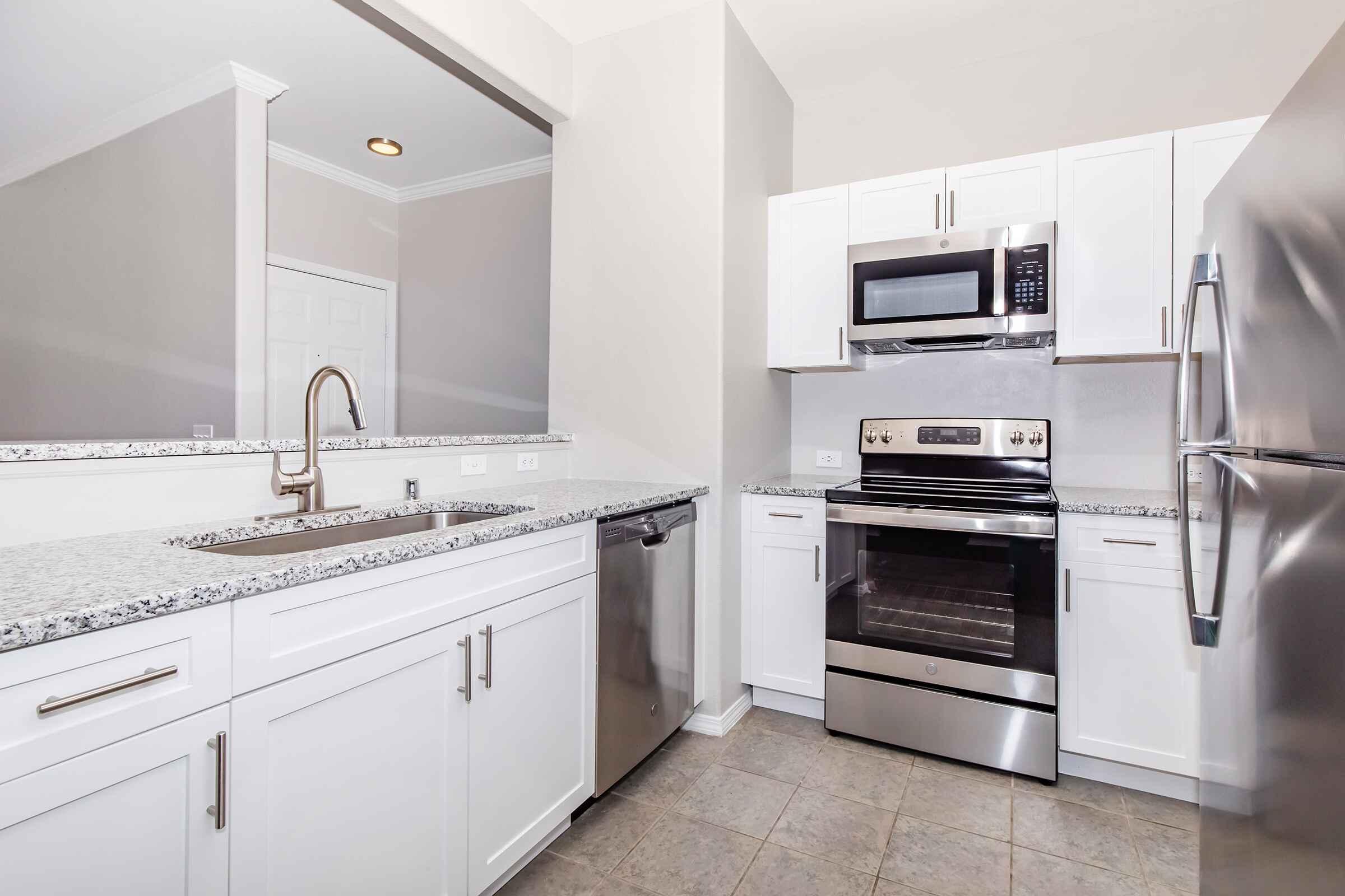 Modern kitchen featuring white cabinetry, stainless steel appliances including a microwave, oven, and dishwasher, and a granite countertop with an undermount sink. The walls are painted in a light color, and the flooring is a neutral tile.