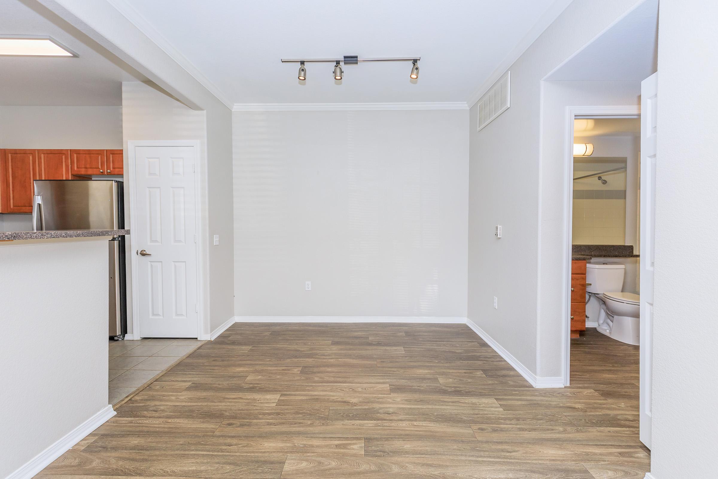 An empty living space featuring light oak-colored laminate flooring. The room includes a kitchen area with wooden cabinets and stainless steel refrigerator, a doorway leading to a bathroom, and natural light coming through the windows. The walls are painted in a neutral color, enhancing the spacious feel.