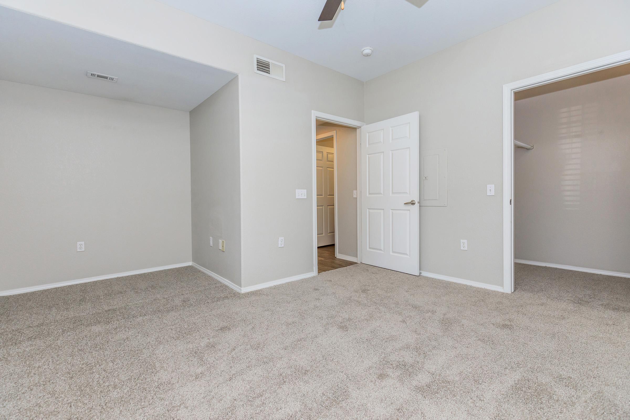 Empty room with light gray walls and beige carpet. There's a ceiling fan and a doorway leading to a closet on the left, with another door slightly ajar in the background. The space appears clean and uncluttered, emphasizing a minimalist style.