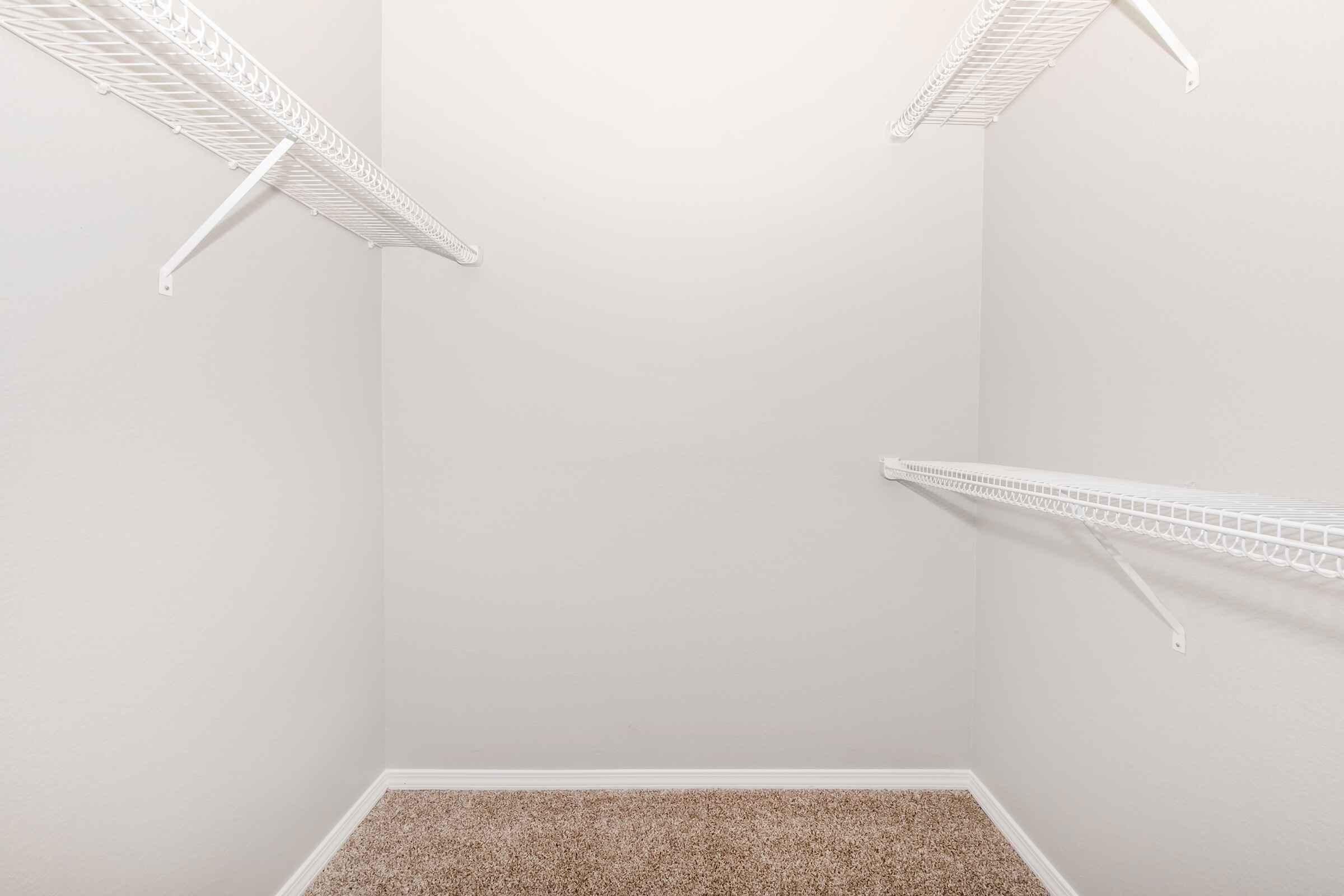 An empty walk-in closet with light gray walls and beige carpet. It features white wire shelving on two sides, providing storage options. The overall space has a clean and minimalistic appearance, emphasizing the openness of the closet.
