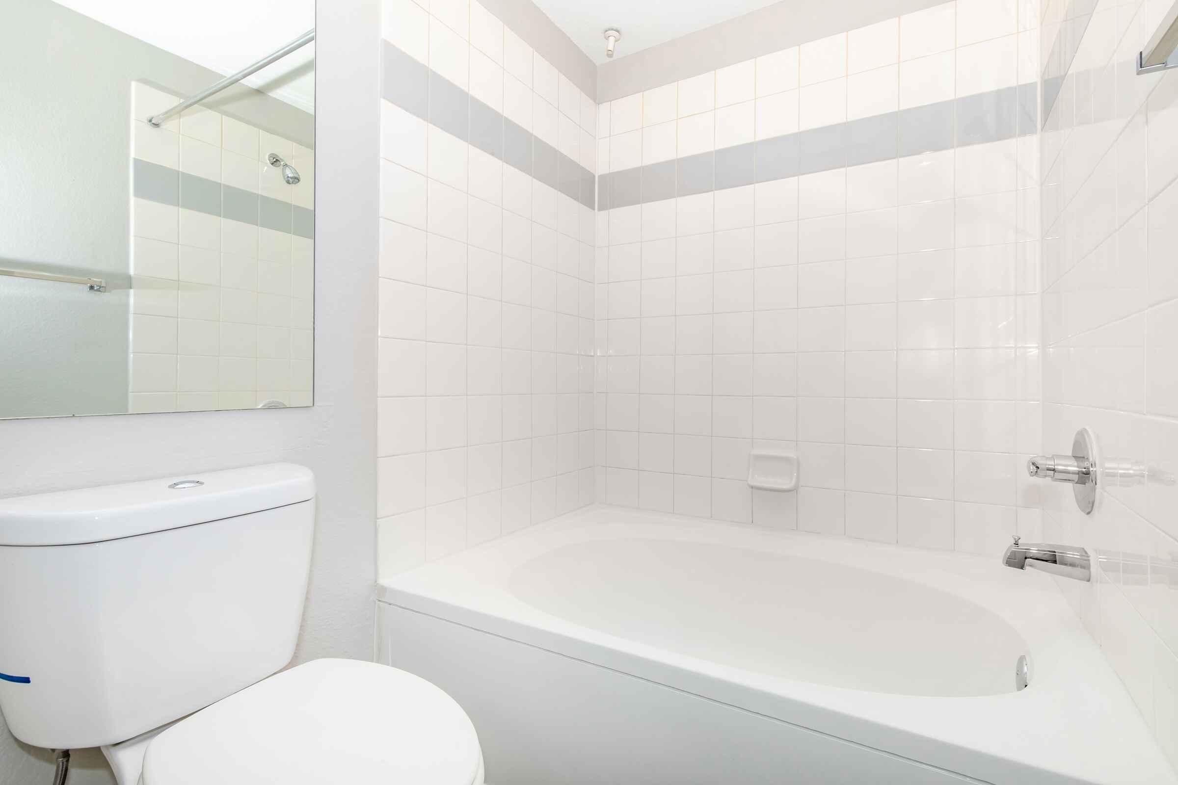 Clean bathroom featuring a white tiled shower and bathtub combination, a modern toilet, and a large mirror above the sink area. The walls are primarily white with a subtle gray accent strip. The overall design is bright and minimalistic.