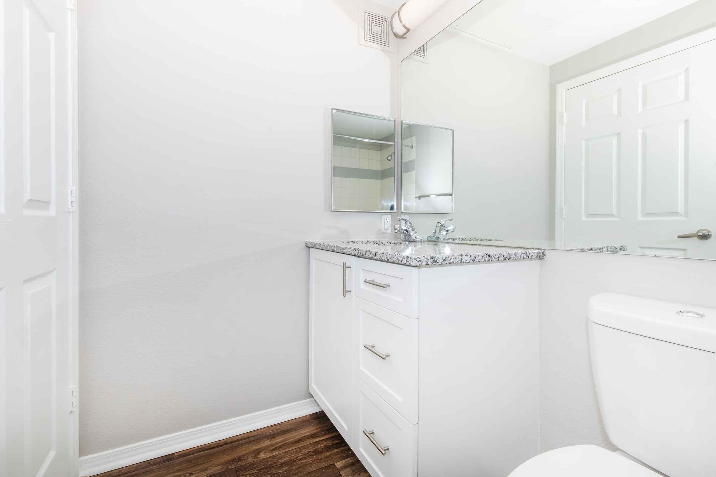 A modern bathroom featuring a white vanity with a granite countertop, a rectangular mirror, a chrome faucet, and a toilet. The walls are painted in a light color, and the flooring consists of wooden laminate. The space is well-lit and minimalistic, creating a clean and airy atmosphere.