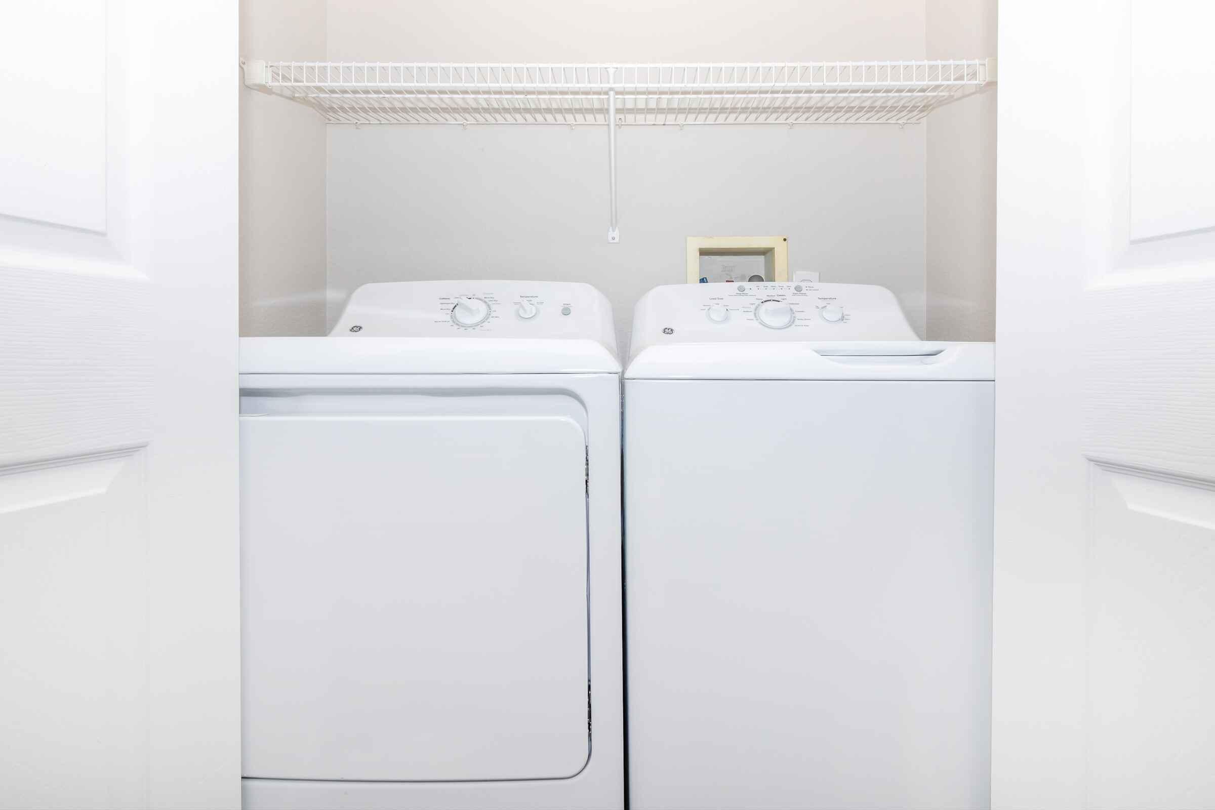 A laundry area featuring a stacked washer and dryer in a closet. The appliances are white and have control dials on the front. Above them, there is a shelf for storage, and the walls are painted light-colored, creating a clean and organized space.