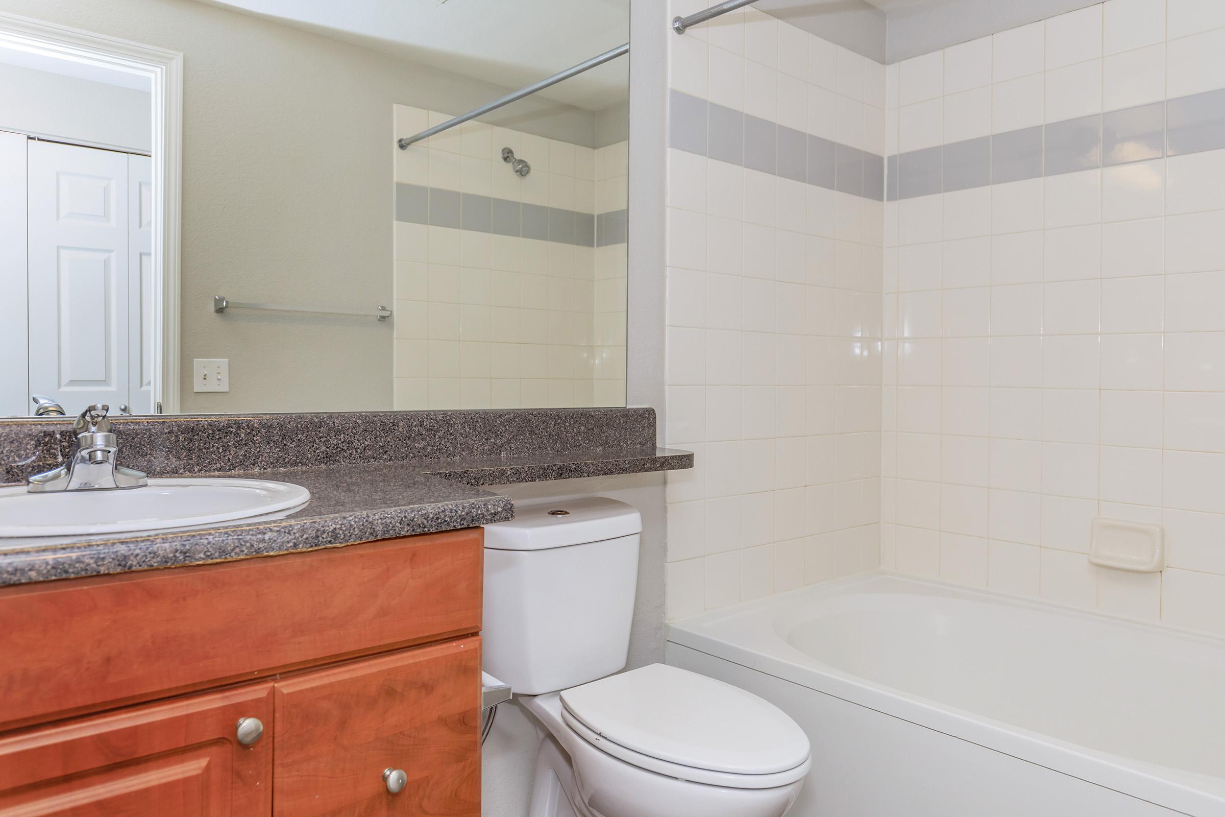 A modern bathroom featuring a white bathtub, a toilet, and a sink with a dark granite countertop. The walls are tiled in light colors with a gray stripe, and there is a mirror above the sink. In the background, a door leads to another room. The overall design is clean and contemporary.