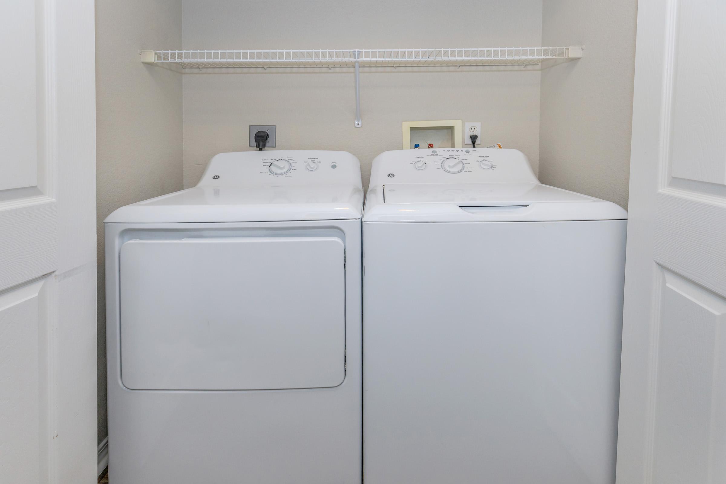 A white washer and dryer side by side in a laundry closet. The appliances are positioned against a light-colored wall, with a shelf above them. The space is well-lit and features two white doors leading to the closet.