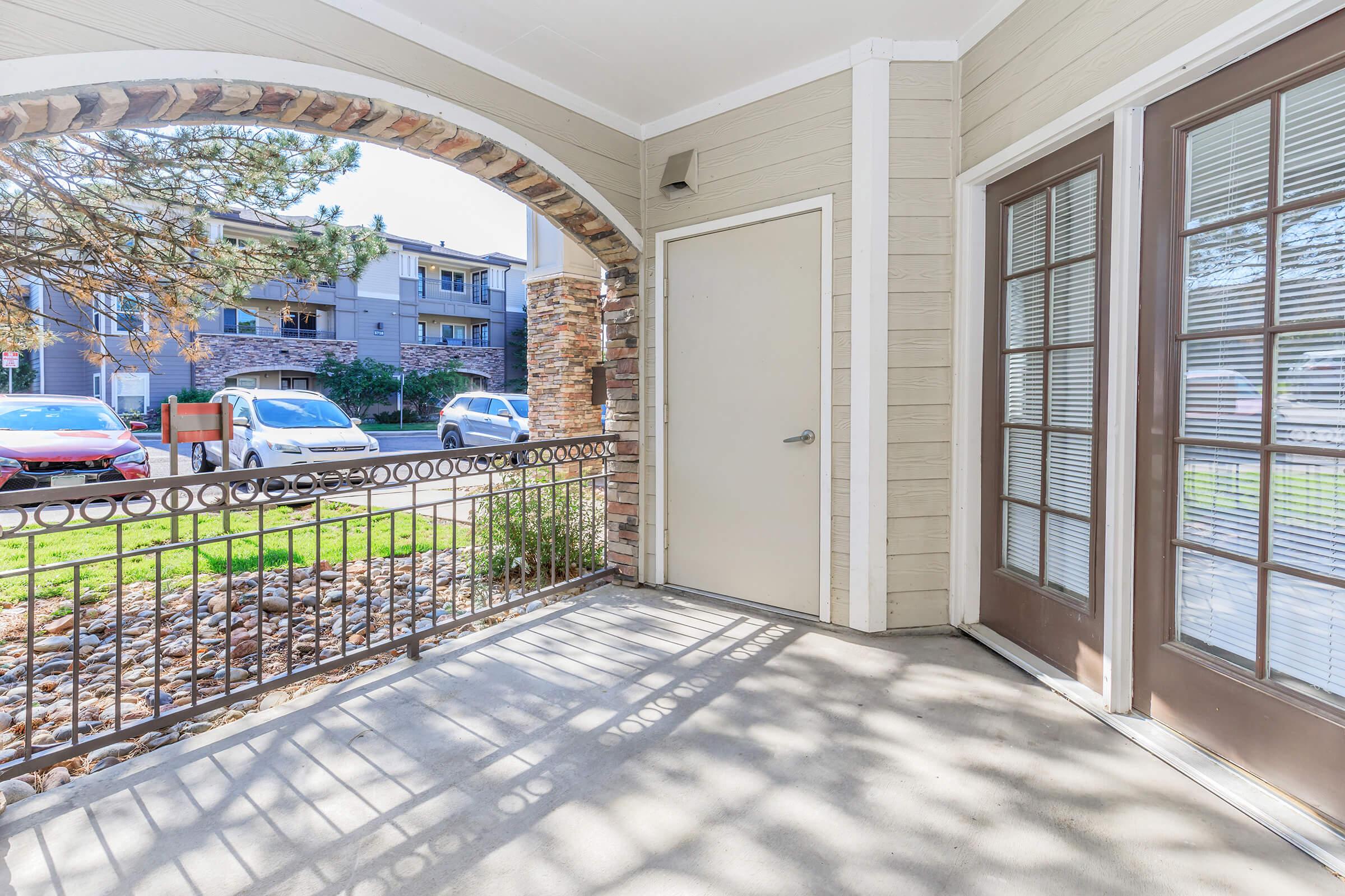 A covered porch with an arched entryway, featuring a door on the left and large windows on the right. The ground is concrete and landscaped with small stones. In the background, there are parked cars and apartments visible across the street. Natural light casts shadows on the surface.