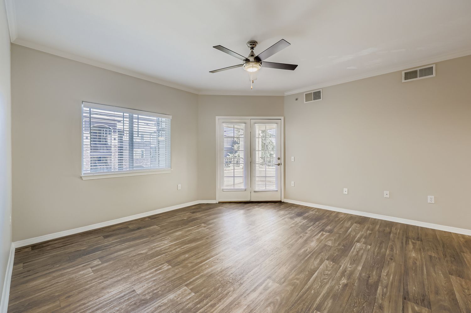 A spacious, empty room with light brown wooden flooring, a ceiling fan, and a large window providing natural light. There are sliding glass doors leading to a patio area, and the walls are painted in a neutral beige color. The overall atmosphere feels bright and open.