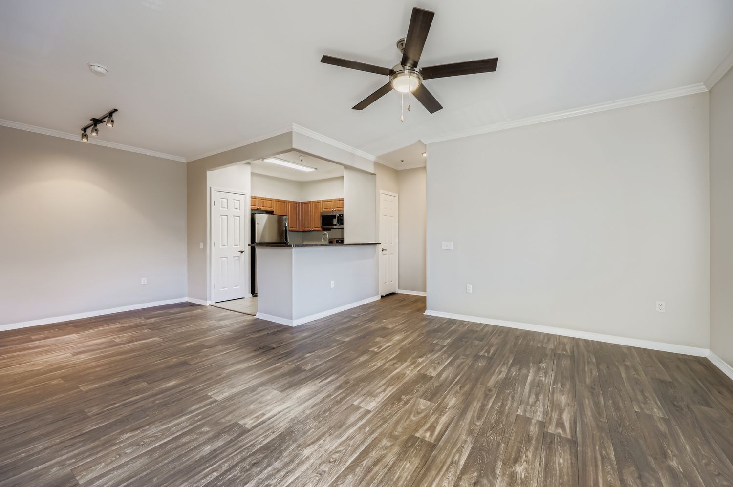 Spacious interior of a room with wooden flooring, a ceiling fan, and light gray walls. An open kitchen area features modern cabinetry and appliances. The room is well-lit, providing a welcoming atmosphere.