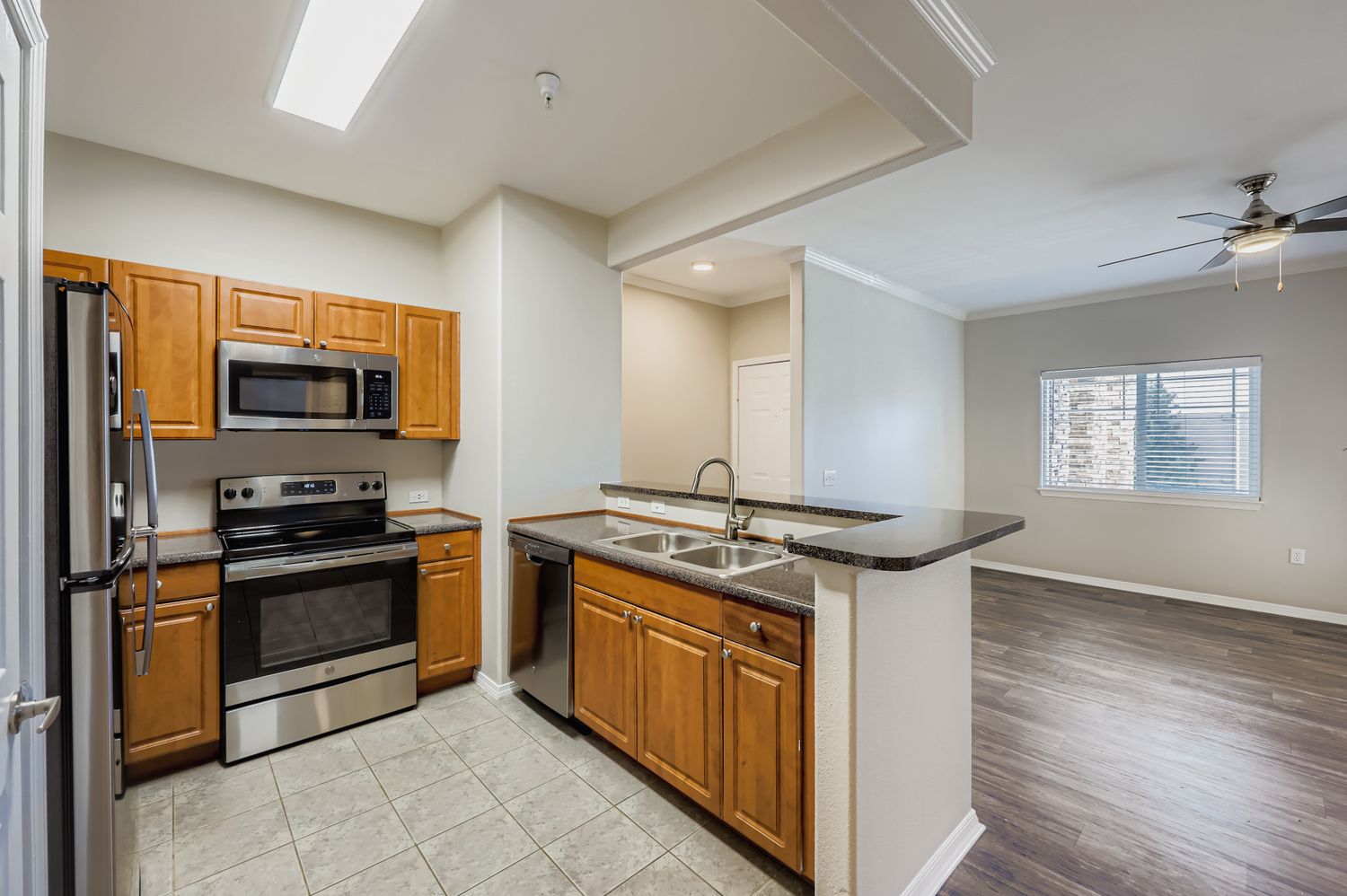 A modern kitchen featuring wooden cabinets, stainless steel appliances including an oven and microwave, a double sink, and a tiled backsplash. The kitchen opens to a spacious living area with laminate flooring and a ceiling fan, illuminated by natural light from a nearby window.