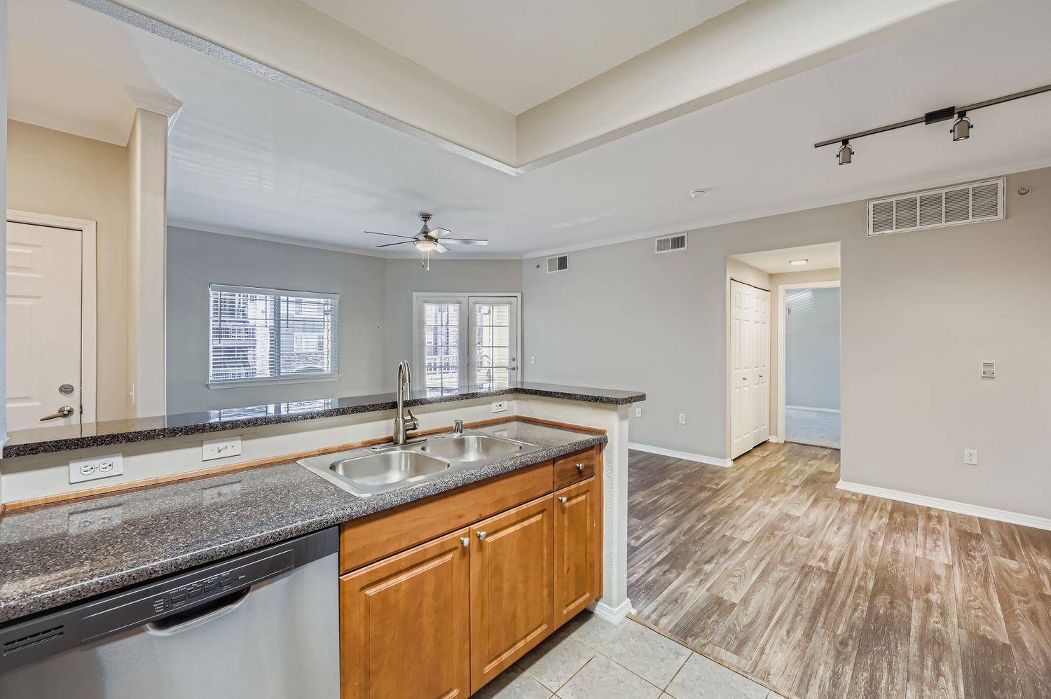 Modern kitchen with dark granite countertops, stainless steel sink, and wooden cabinets. A dishwasher is visible next to the sink. The open layout leads to a living area with light-colored flooring. Natural light comes through windows, and a ceiling fan is mounted above. A hallway is seen in the background.