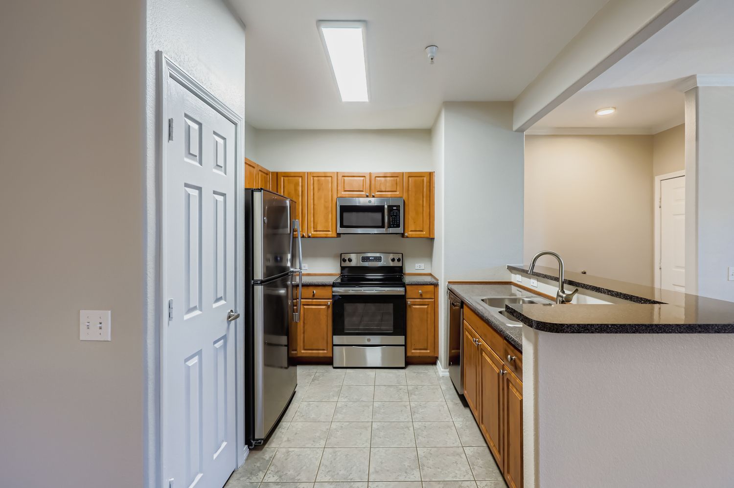 Modern kitchen featuring wooden cabinets, stainless steel appliances including a refrigerator, oven, and microwave, along with a dark countertop. Bright lighting from a ceiling fixture illuminates the space, which includes tiled flooring and a partial view of an adjacent room.