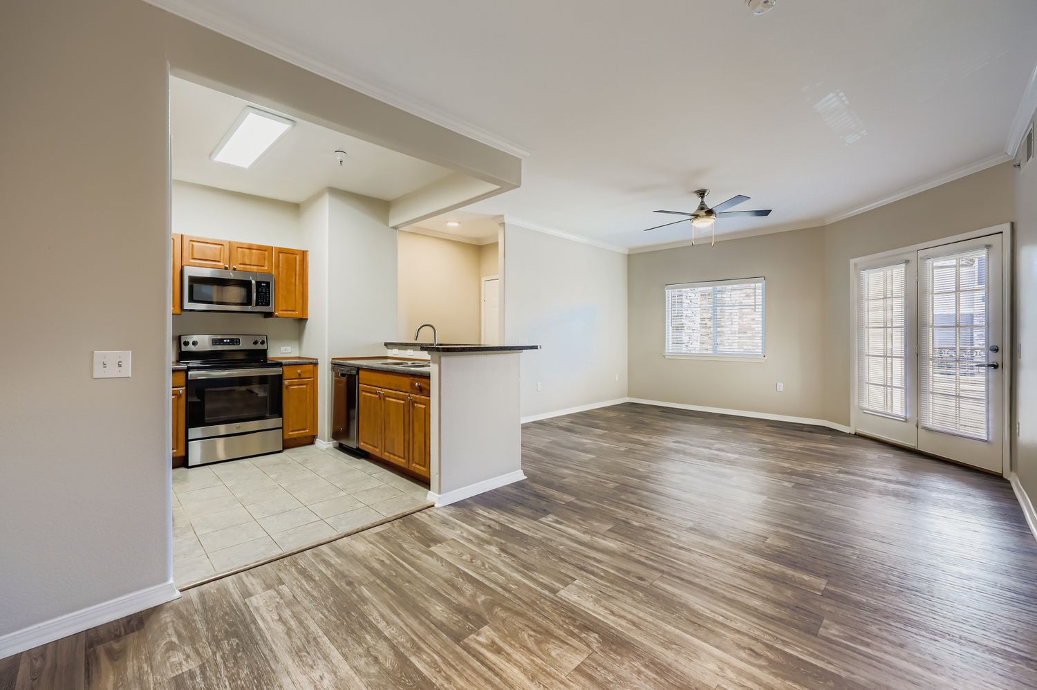 Spacious living area with hardwood floors, featuring a ceiling fan and large windows. To the left, a kitchen with wooden cabinets, stainless steel appliances, and tile flooring is visible. The room opens to a patio door leading outside, creating a bright, inviting atmosphere.
