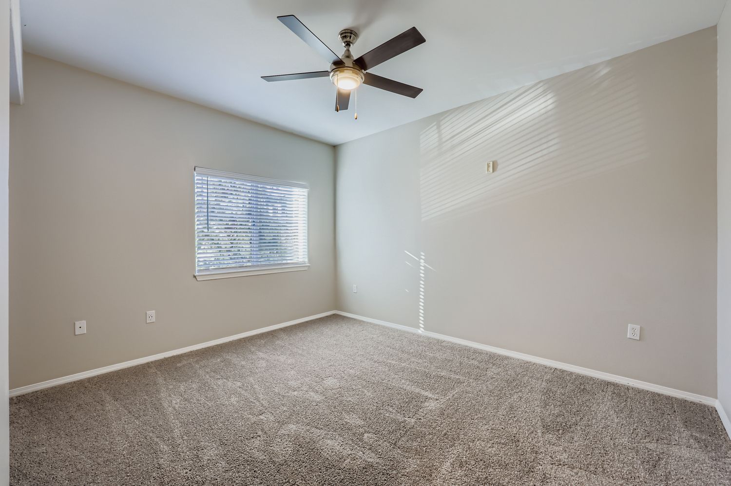 A bright and airy room with light beige walls and plush carpet. A window with blinds allows natural light to filter in, and a ceiling fan is mounted in the center. The space is empty, creating a feeling of openness and potential for personalization.