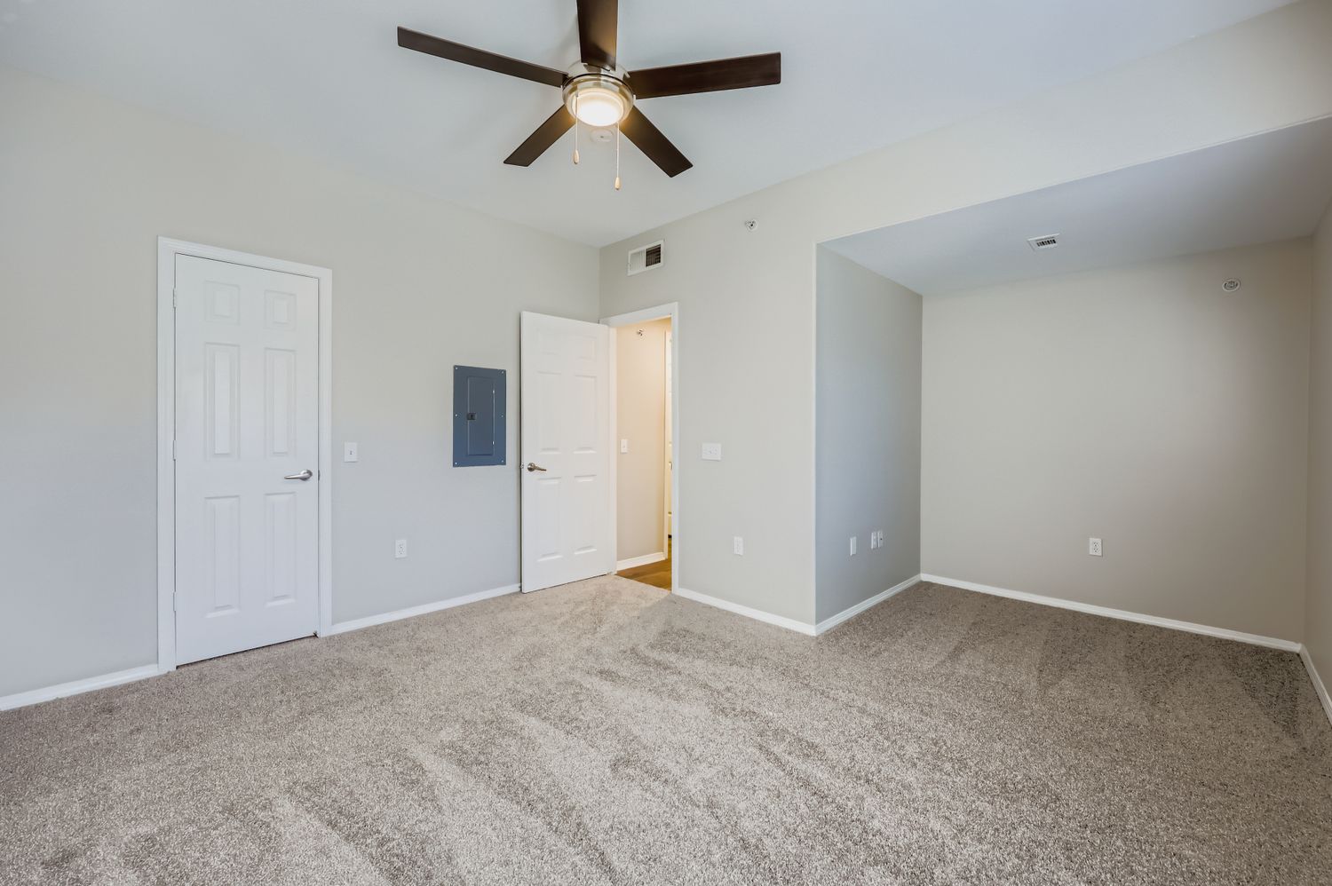 A spacious room with light gray walls and plush carpet. It features a ceiling fan, a door leading to another area, and a closet. There is a small nook on one wall and a wall-mounted electrical panel. The room is well-lit with natural light and has a clean, modern aesthetic.