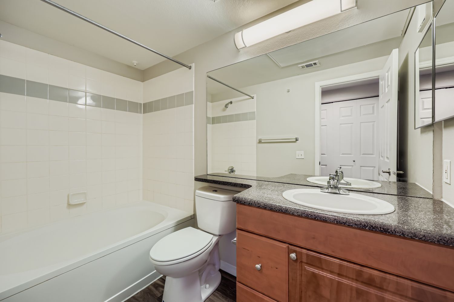 A clean and modern bathroom featuring a bathtub with a shower, a white toilet, and a double sink vanity with dark countertops. The walls are tiled in light colors, and there is a large mirror above the sinks. The overall design is bright and spacious, with a simple yet elegant aesthetic.