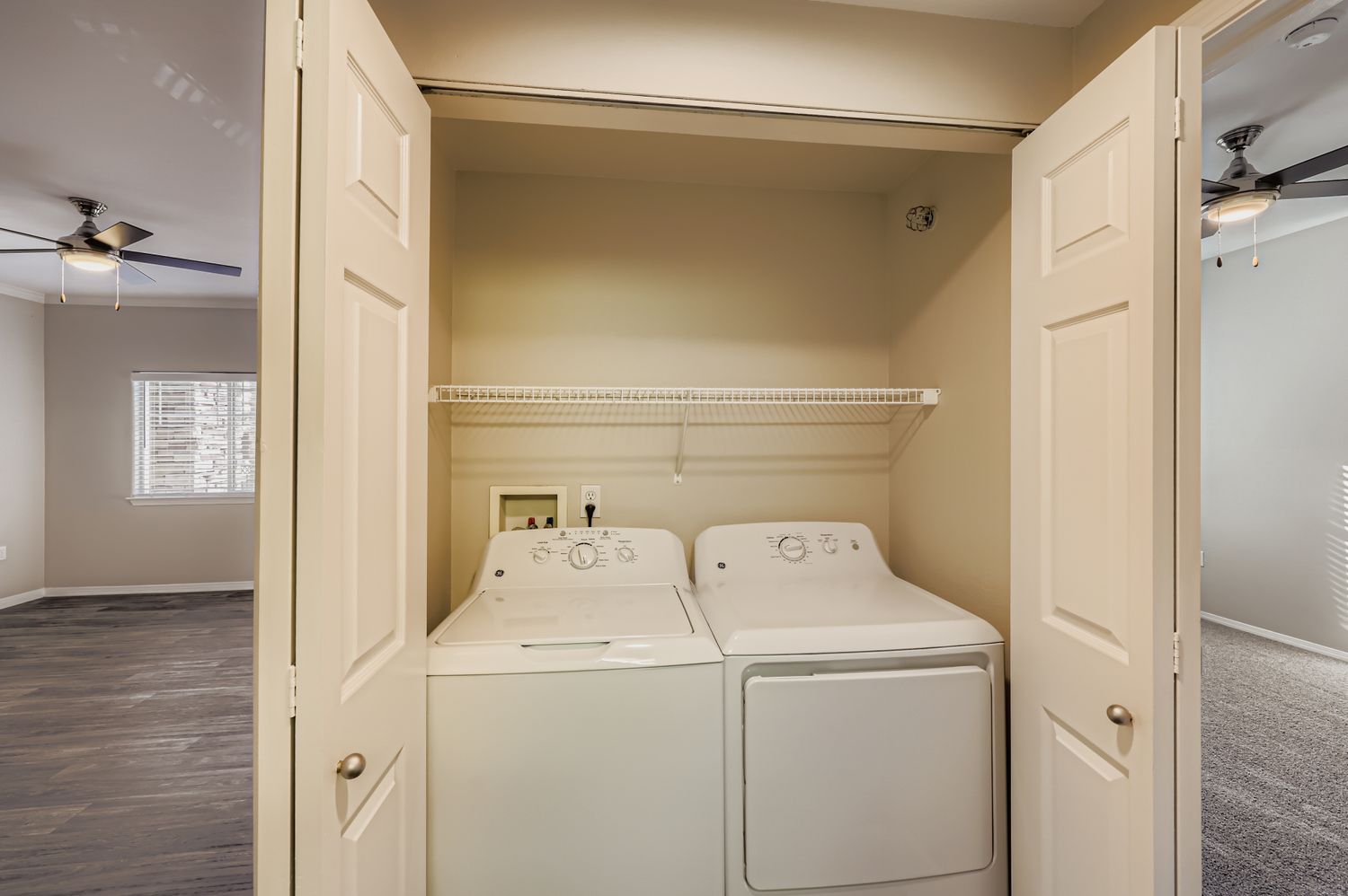 A compact laundry area with a washer and dryer side by side, tucked behind double doors. Above, there is a shelf for storage. The room features neutral walls and is adjacent to a living space with ceiling fans and windows, suggesting natural light.