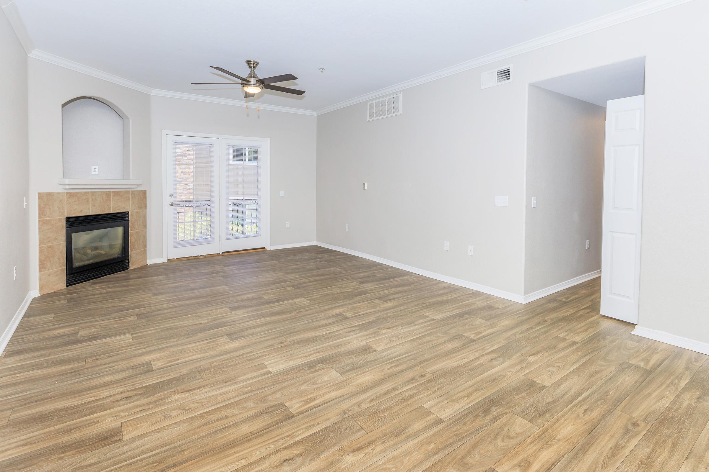 Spacious living room featuring light wood flooring, a ceiling fan, and a cozy fireplace. Large windows allow natural light, and the space includes a doorway leading to another room. Neutral wall colors enhance the open and airy feel of the area.