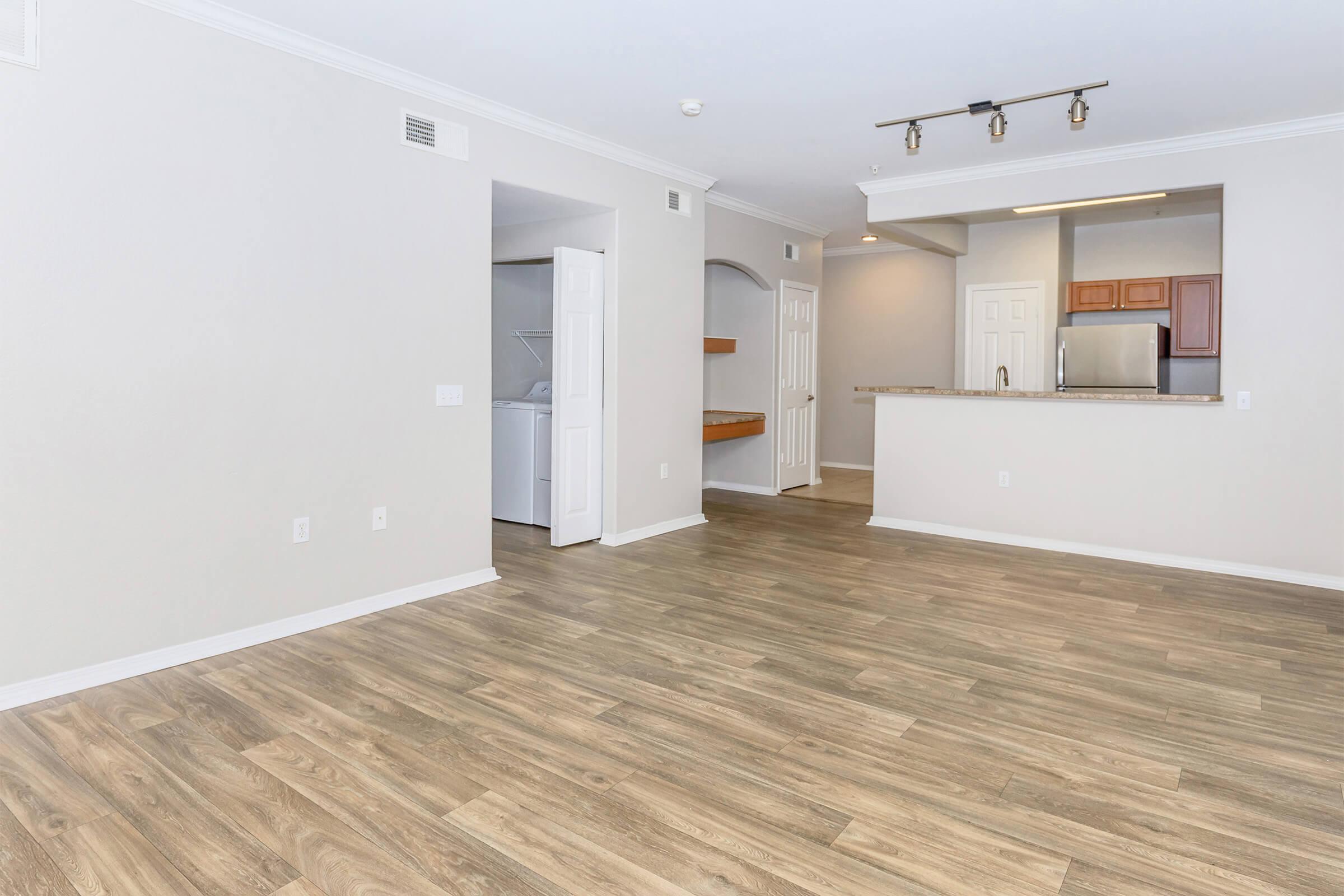 Spacious, empty living area with light-colored walls and wood laminate flooring. A kitchen bar area is visible in the background, along with a laundry nook and a doorway. The space is well-lit, featuring modern fixtures and an open layout. Ideal for a cozy, modern living experience.