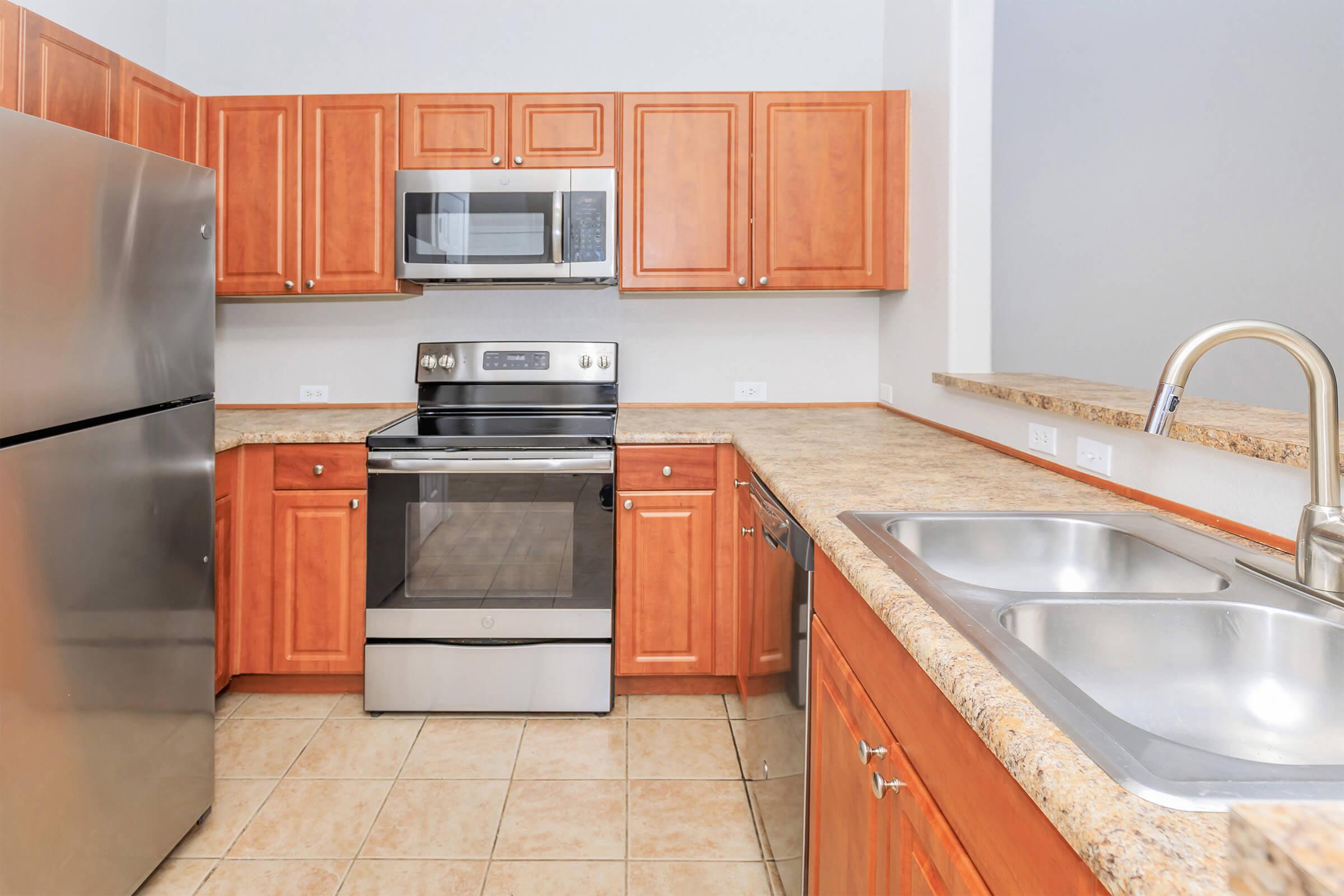 Modern kitchen featuring wooden cabinets, stainless steel refrigerator, microwave, and oven. The countertop is made of granite, and there's a double sink with a sleek faucet. The floor is tiled, and the walls are painted a light color, creating a bright and inviting atmosphere.