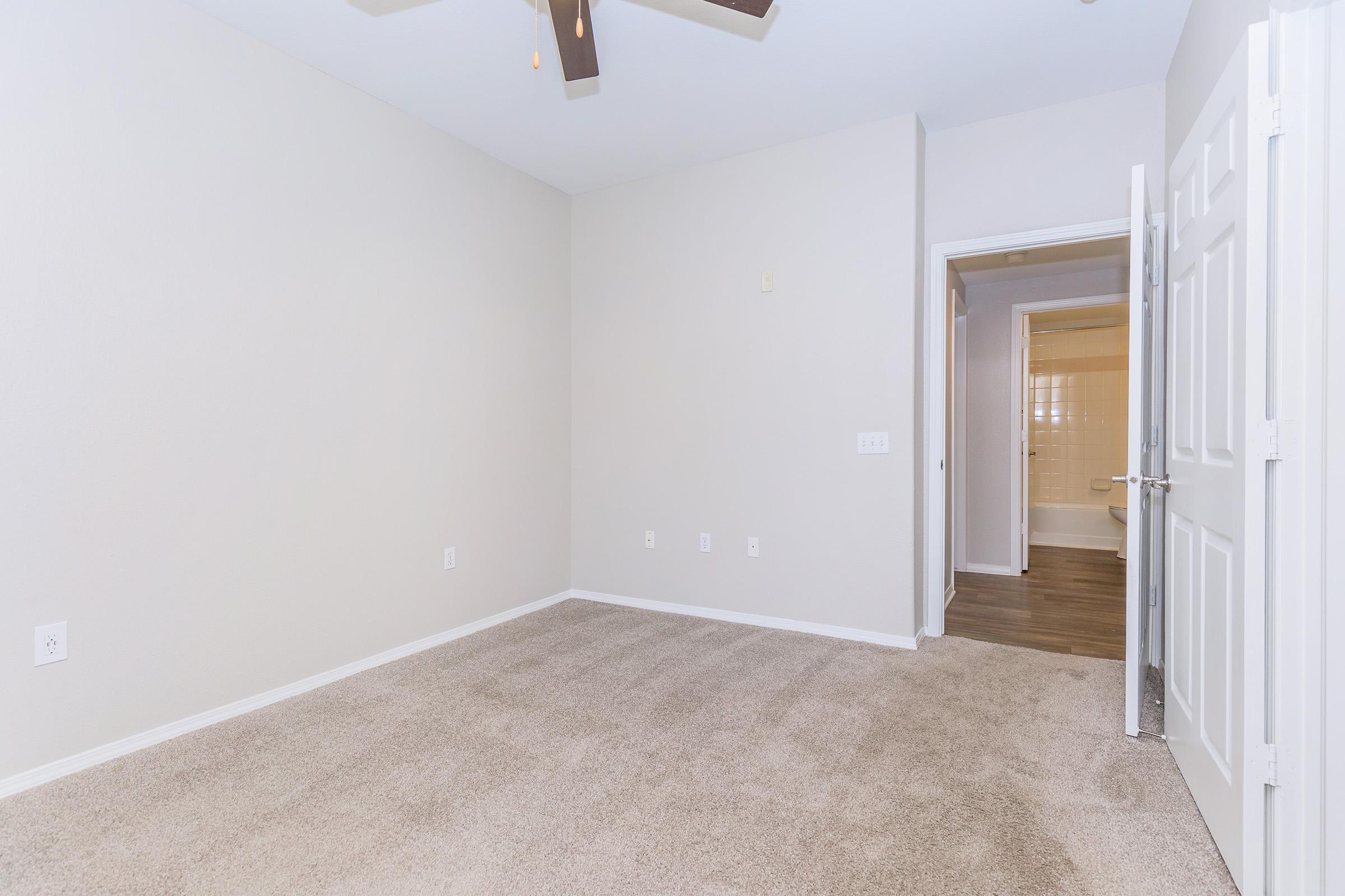 Empty room with light-colored walls and beige carpet. A ceiling fan is mounted on the ceiling. A doorway leads to a bathroom, visible in the background. The space is well-lit and clean, creating a neutral atmosphere suitable for personal decoration.