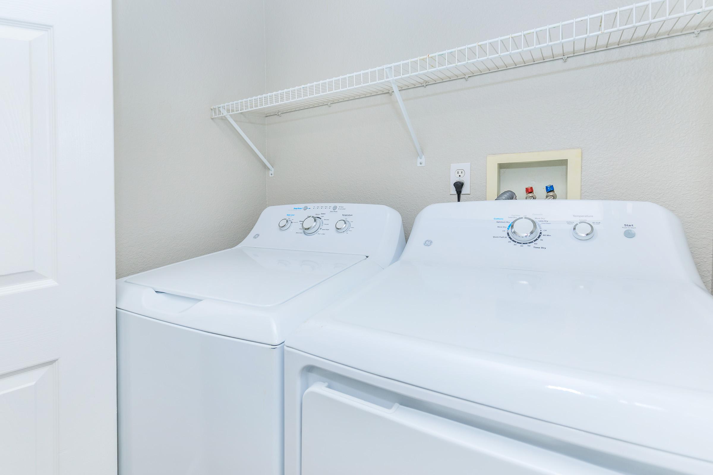 A clean laundry room featuring a white washing machine and a matching dryer side by side. Above them, a wire shelf is mounted on the wall, and a small framed item or outlet can be seen in the background. The walls are light-colored, creating a bright and tidy atmosphere.