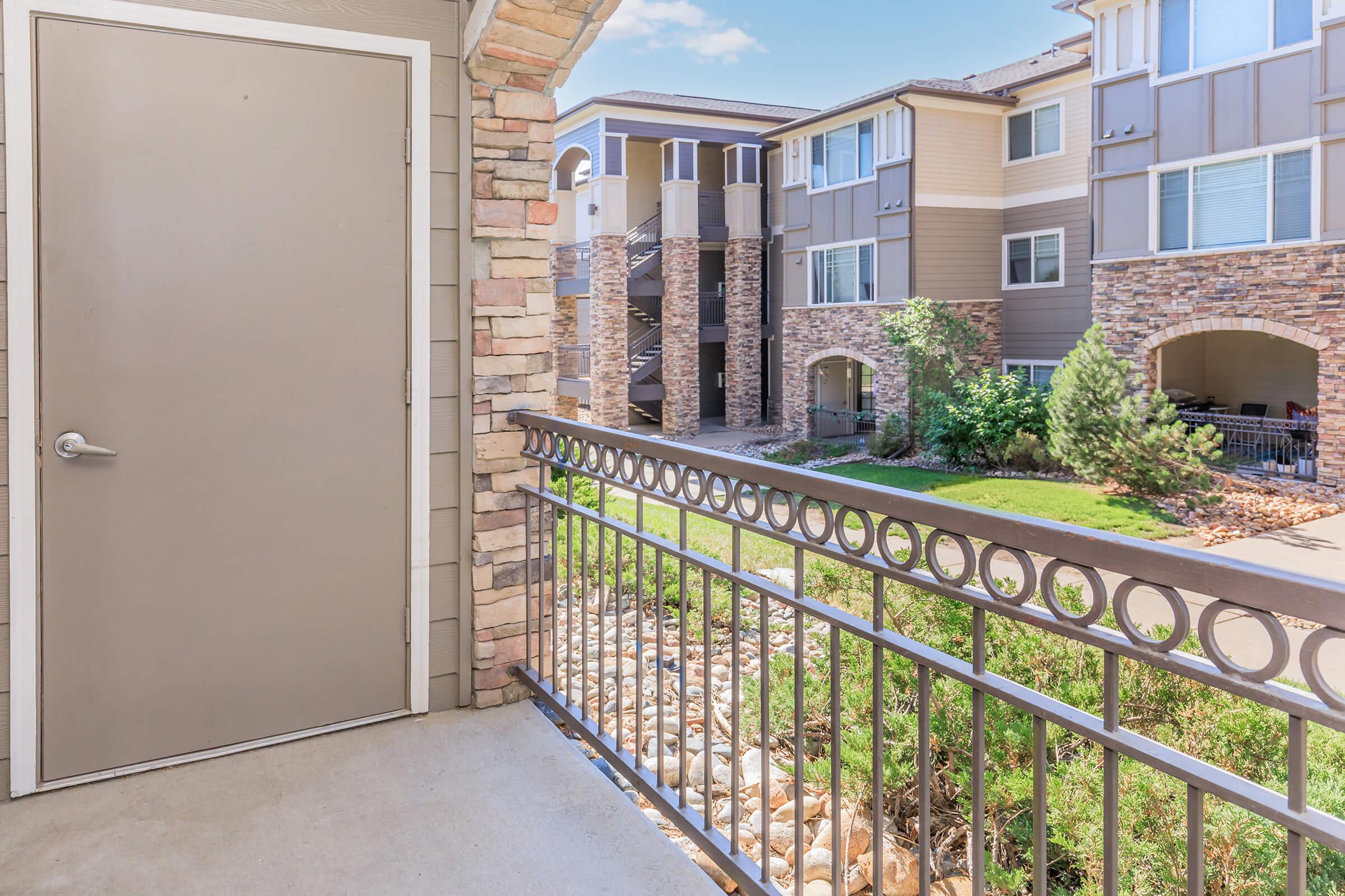 View from a balcony showing a spacious outdoor area with a railing. In the background, multiple apartment buildings are visible, featuring stone accents and greenery. The scene is bright and inviting, with a clear sky overhead.