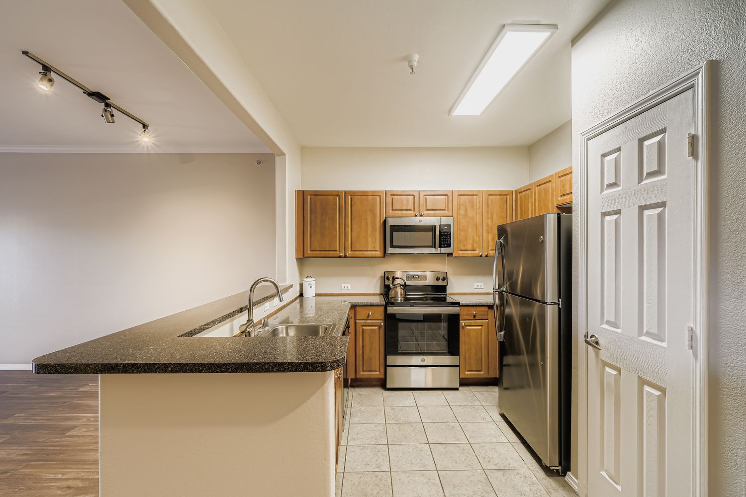 Modern kitchen featuring wooden cabinetry, stainless steel appliances including a microwave and refrigerator, a double sink, and a granite countertop. Soft lighting and a doorway leading to another room are visible, along with tiled flooring.