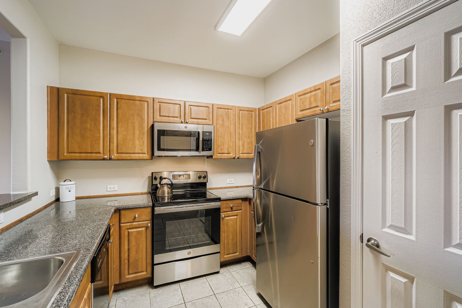 Modern kitchen featuring wooden cabinets, a stainless steel refrigerator, a microwave, and an electric stove with an oven. Countertops are gray with a sink positioned near the wall. The kitchen has a neutral-colored wall and a door leading to another area, illuminated by overhead lighting.