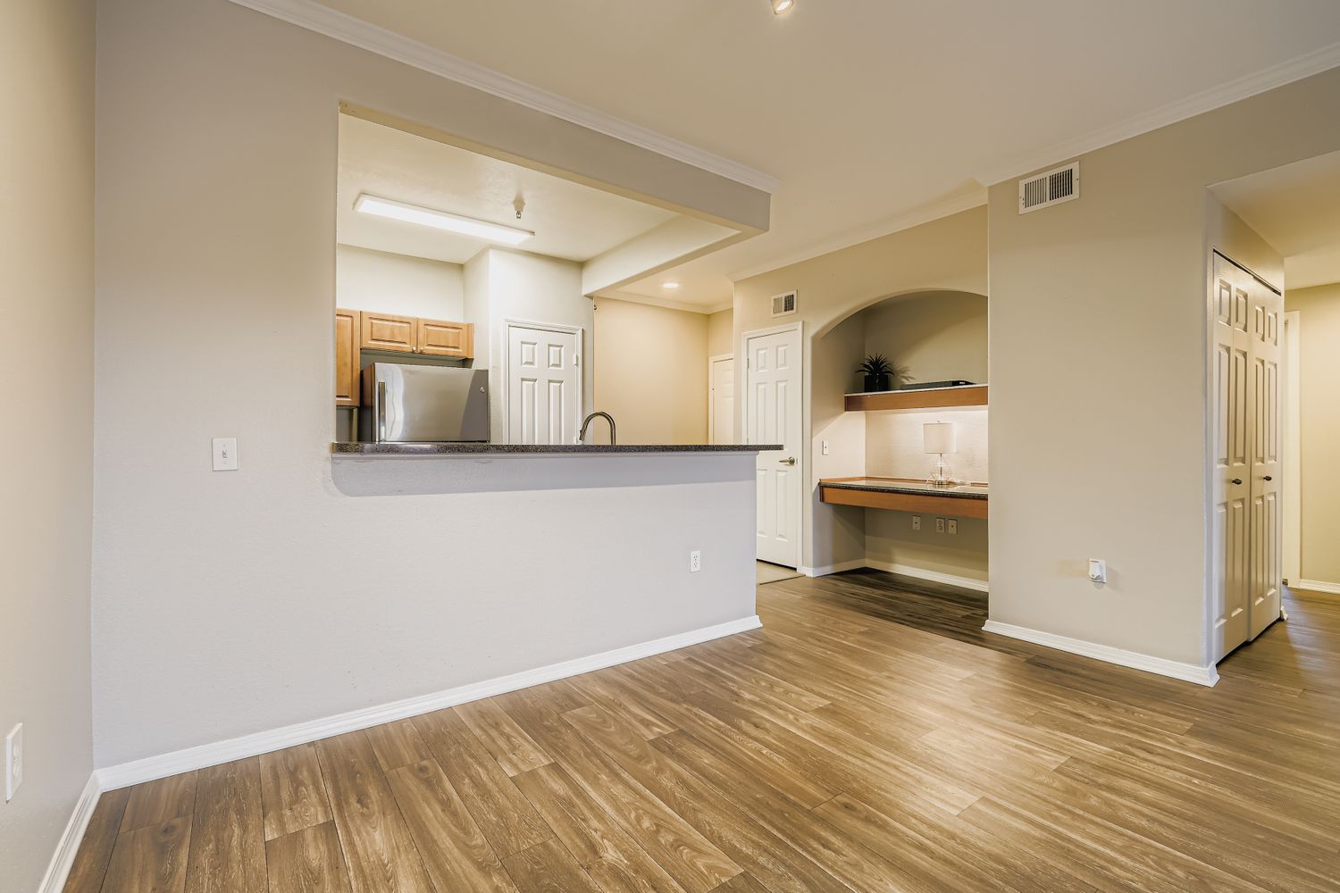 Modern interior shot of a spacious living area featuring hardwood floors, a partially open kitchen with wooden cabinetry and stainless steel appliances. A small nook with a countertop and built-in shelves is visible, along with white doors leading to closets. The walls are painted in neutral tones, creating a warm ambiance.