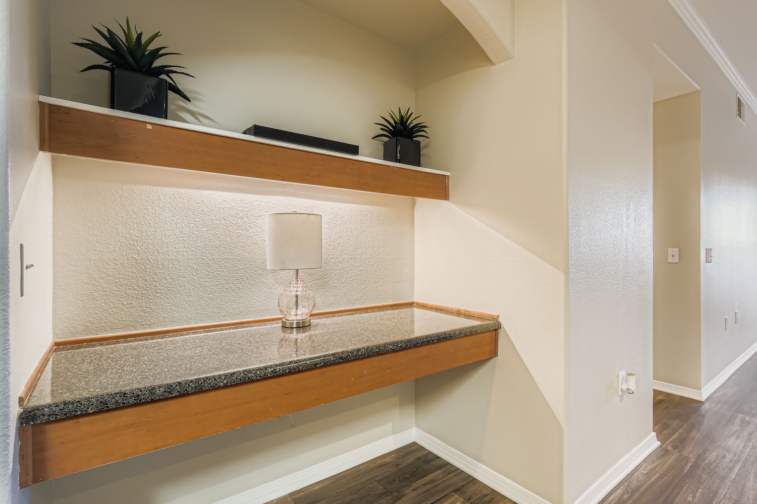A small interior space featuring a minimalist desk with a granite surface, complemented by two potted plants on a wooden shelf above. A clear glass lamp sits on the desk, providing soft lighting. The wall is painted a light color, and the flooring appears wooden, creating a contemporary and inviting atmosphere.