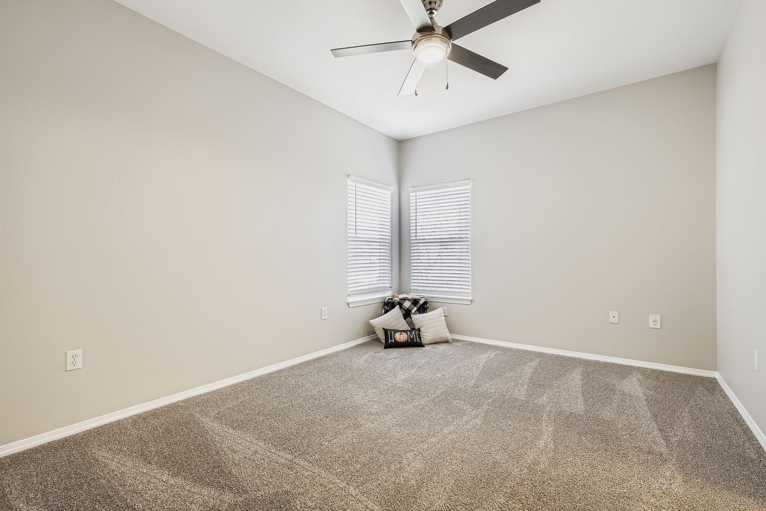 A clean, empty room with light gray walls and a ceiling fan. There are two windows with white blinds letting in natural light. A cozy corner features a few pillows on the carpeted floor, creating a simple, inviting atmosphere.
