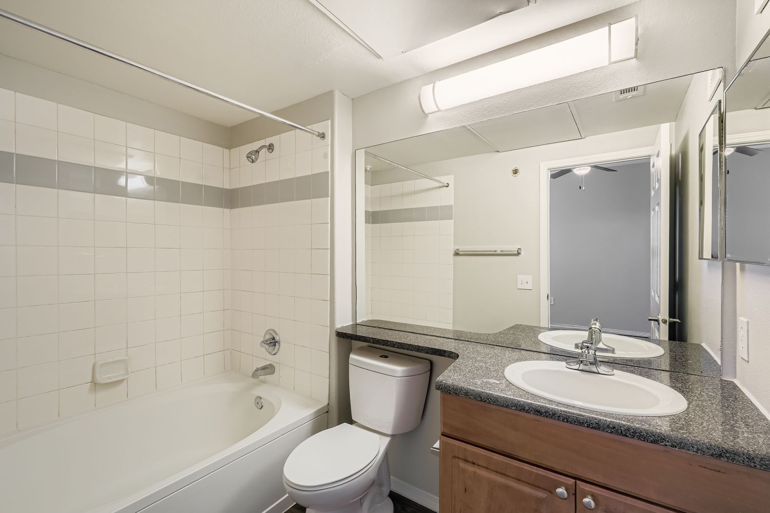 A modern bathroom featuring a bathtub with a shower, a double vanity with two sinks, and a large mirror. The walls are tiled in a light color with gray accents. There is a single light fixture above the mirror and the decor is simple and clean, creating a spacious feel.