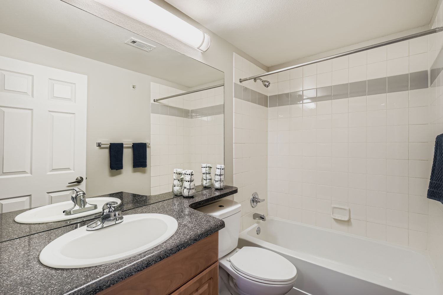 Modern bathroom featuring a bathtub, a toilet, and a sink with a dark countertop. A large mirror reflects the space, and there are neatly arranged towels on a rack. White tiles line the shower area, complemented by a light overhead fixture. The overall design is clean and functional.