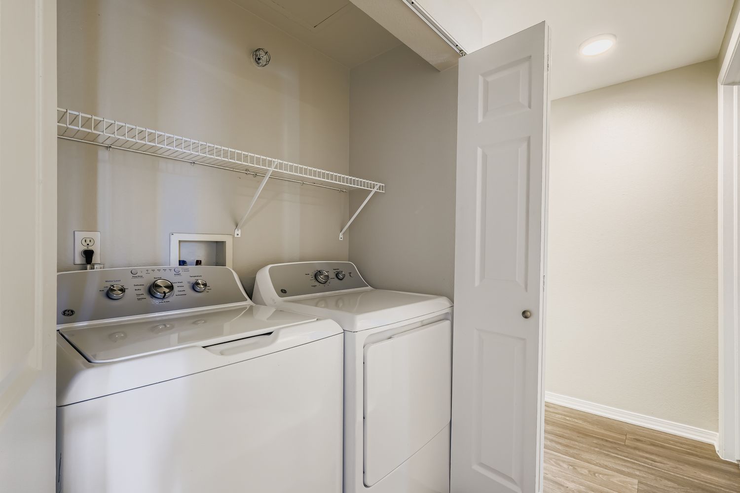 A clean laundry room featuring a stacked washer and dryer set against a light-colored wall. There is a white wire shelf above the appliances for storage, and a door partially open leading to another space. The floor is wood-look, adding a modern touch to the area.