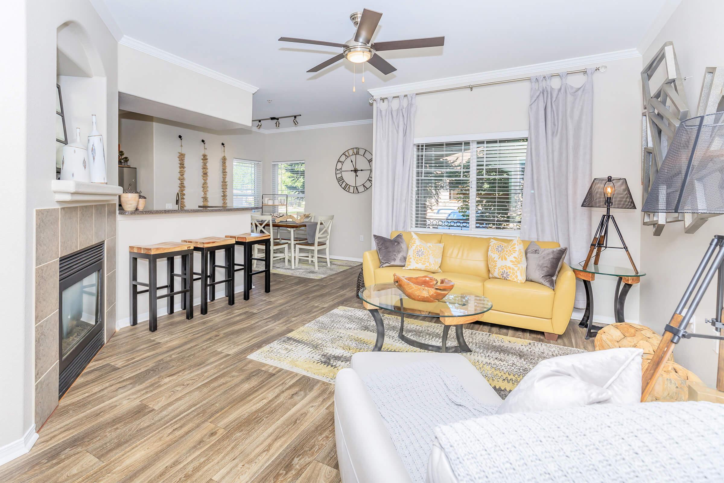 A cozy living room featuring a yellow sofa, glass coffee table, and modern decor. There's a stone fireplace, a dining area in the background with a clock on the wall, and large windows with natural light. The space has a warm and inviting atmosphere with wooden floors and stylish accents.