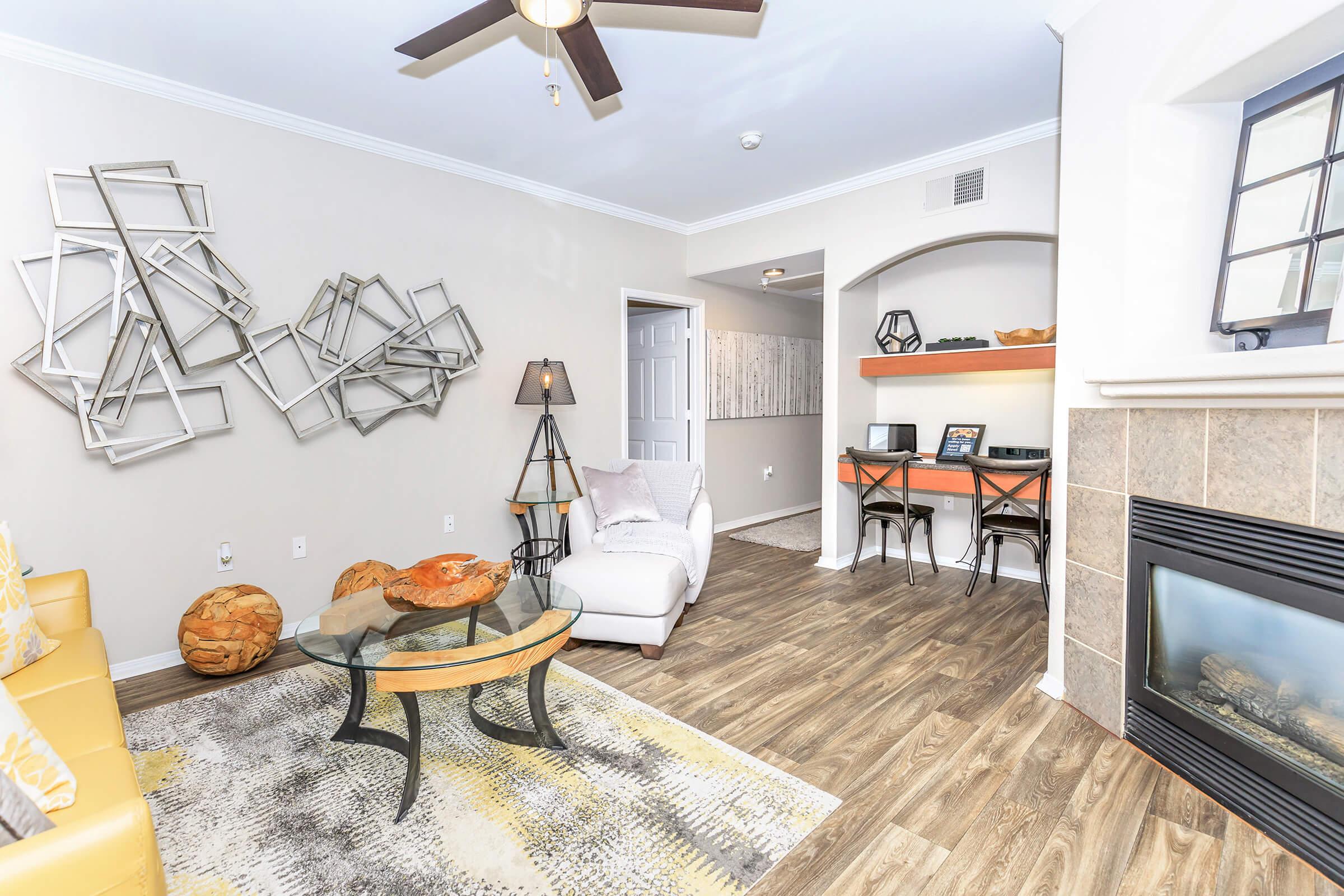 A cozy living room featuring a yellow sofa, a white armchair, and a coffee table on a patterned area rug. Wall art with geometric designs hangs above. A fireplace and a small desk setup are visible, creating a warm and inviting atmosphere. Natural light brightens the space.