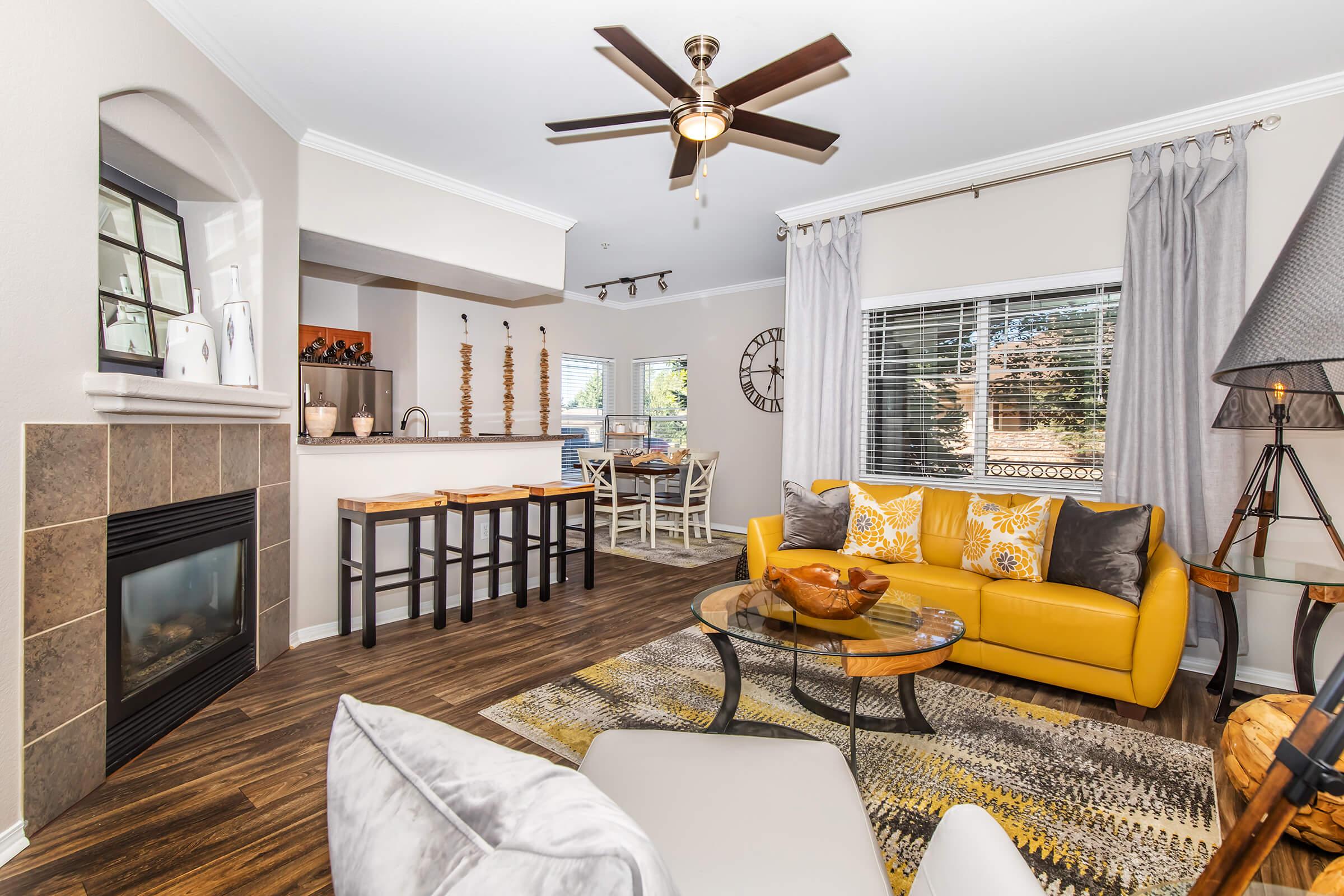 A cozy living room featuring a bright yellow sofa with decorative pillows, a round coffee table, a textured area rug, and a stylish ceiling fan. The space includes a fireplace, large windows with sheer curtains, and a dining area visible in the background. Warm, inviting decor creates a welcoming atmosphere.
