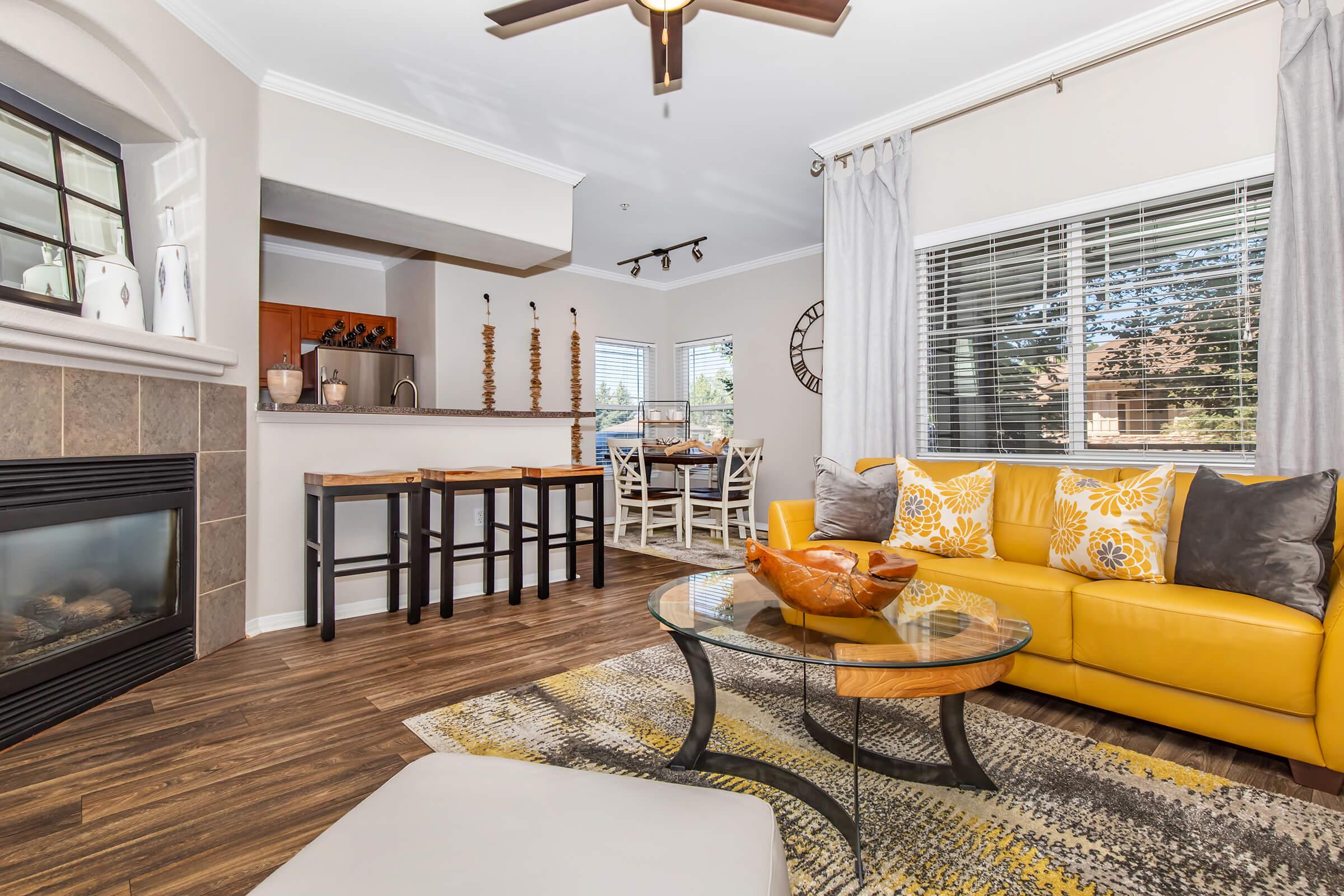 A stylish living room featuring a bright yellow sofa adorned with decorative pillows, a glass coffee table with an orange centerpiece, and a cozy fireplace. The decor includes modern furnishings, a dining area visible in the background, and large windows allowing natural light to fill the space.