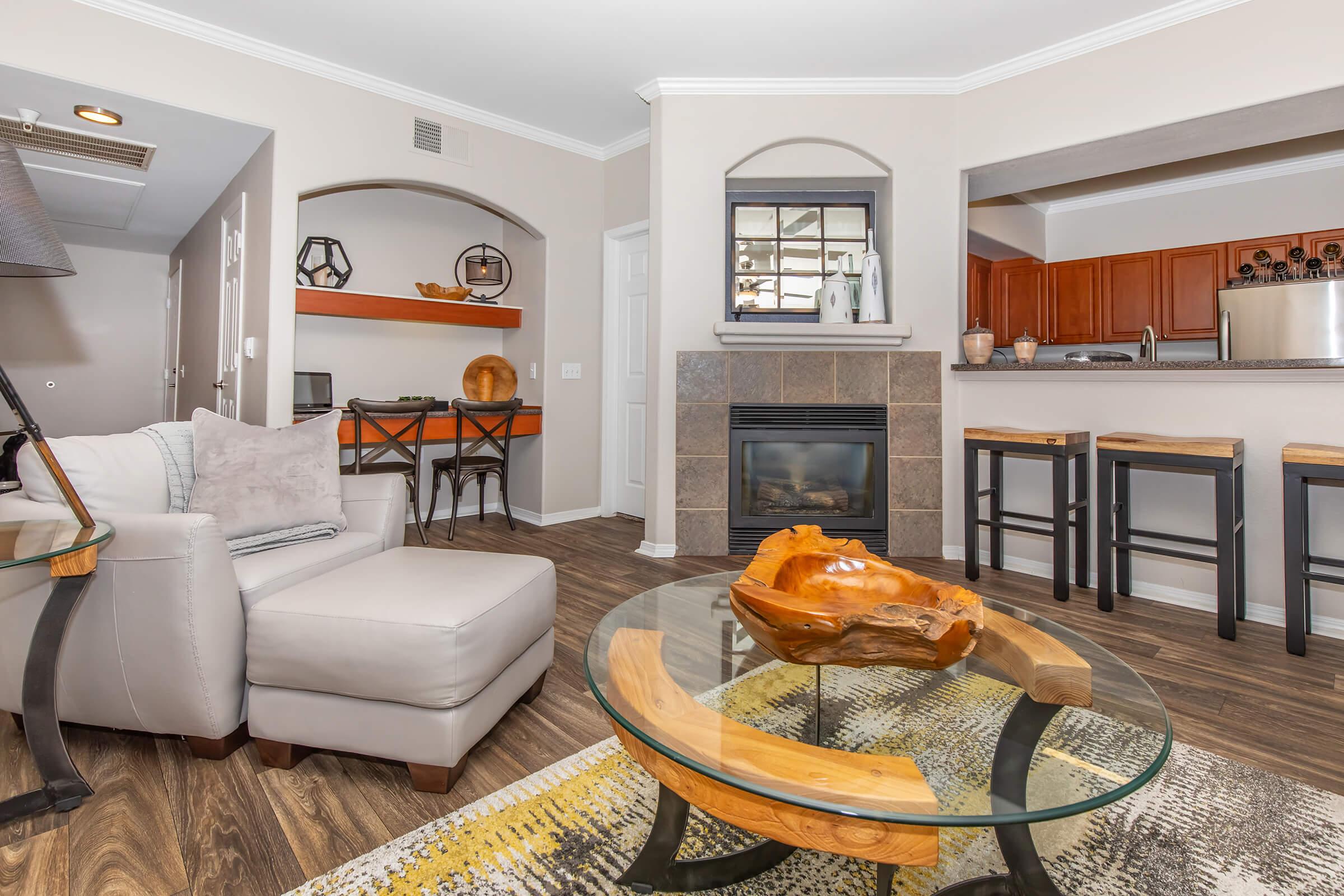 A cozy living room featuring a light-colored sofa, a round glass coffee table with a wooden centerpiece, and modern barstools at a kitchen counter. The decor includes wood accents and a fireplace, with neutral tones on the walls and vibrant patterns on the rug, creating a warm and inviting atmosphere.